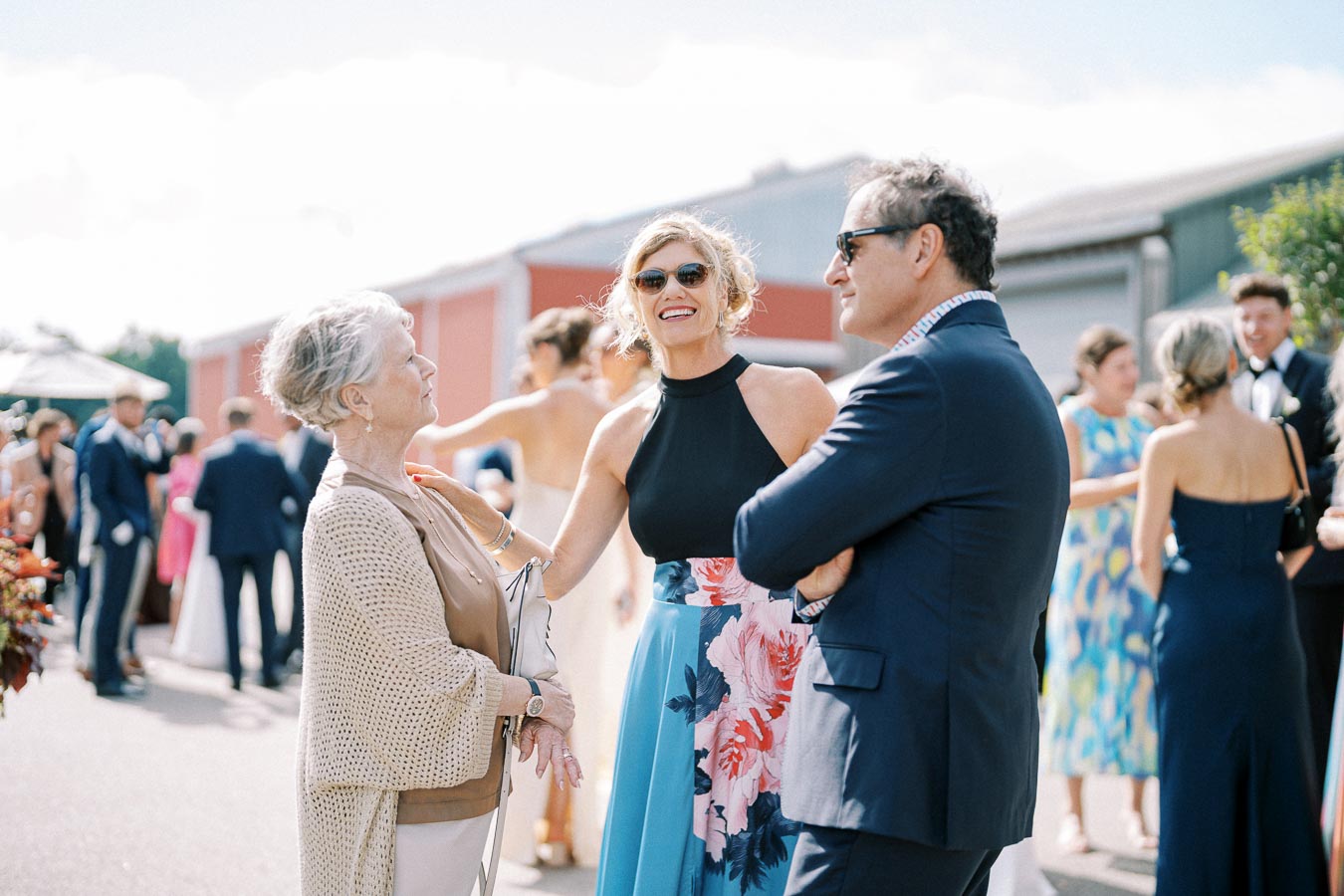 Three people happily conversing at an outdoor event, surrounded by guests in formal attire.