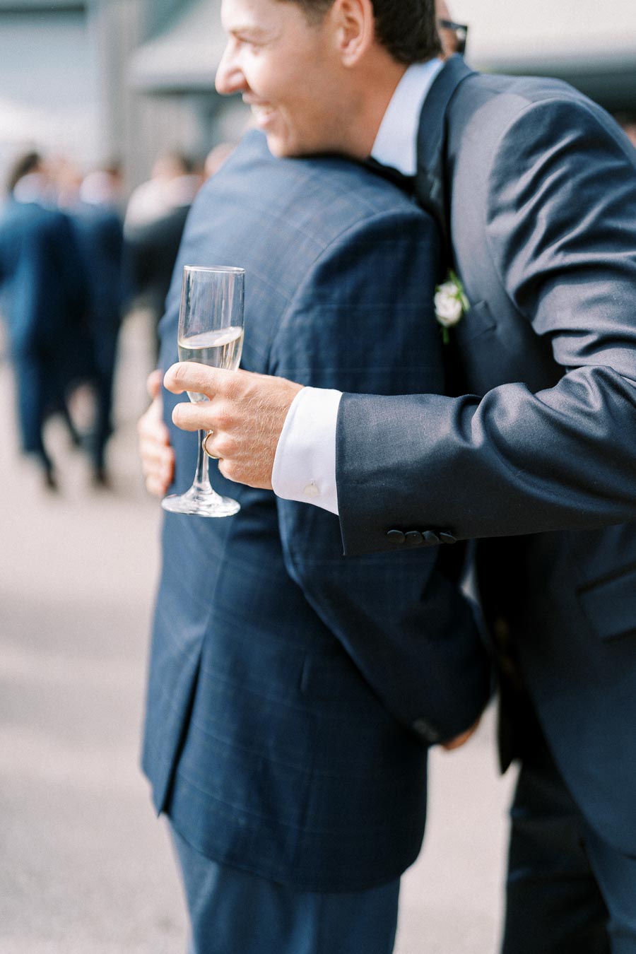 Two men in formal suits embrace at an outdoor event, one holding a champagne glass. The setting appears celebratory, with other attendees visible in the background.
