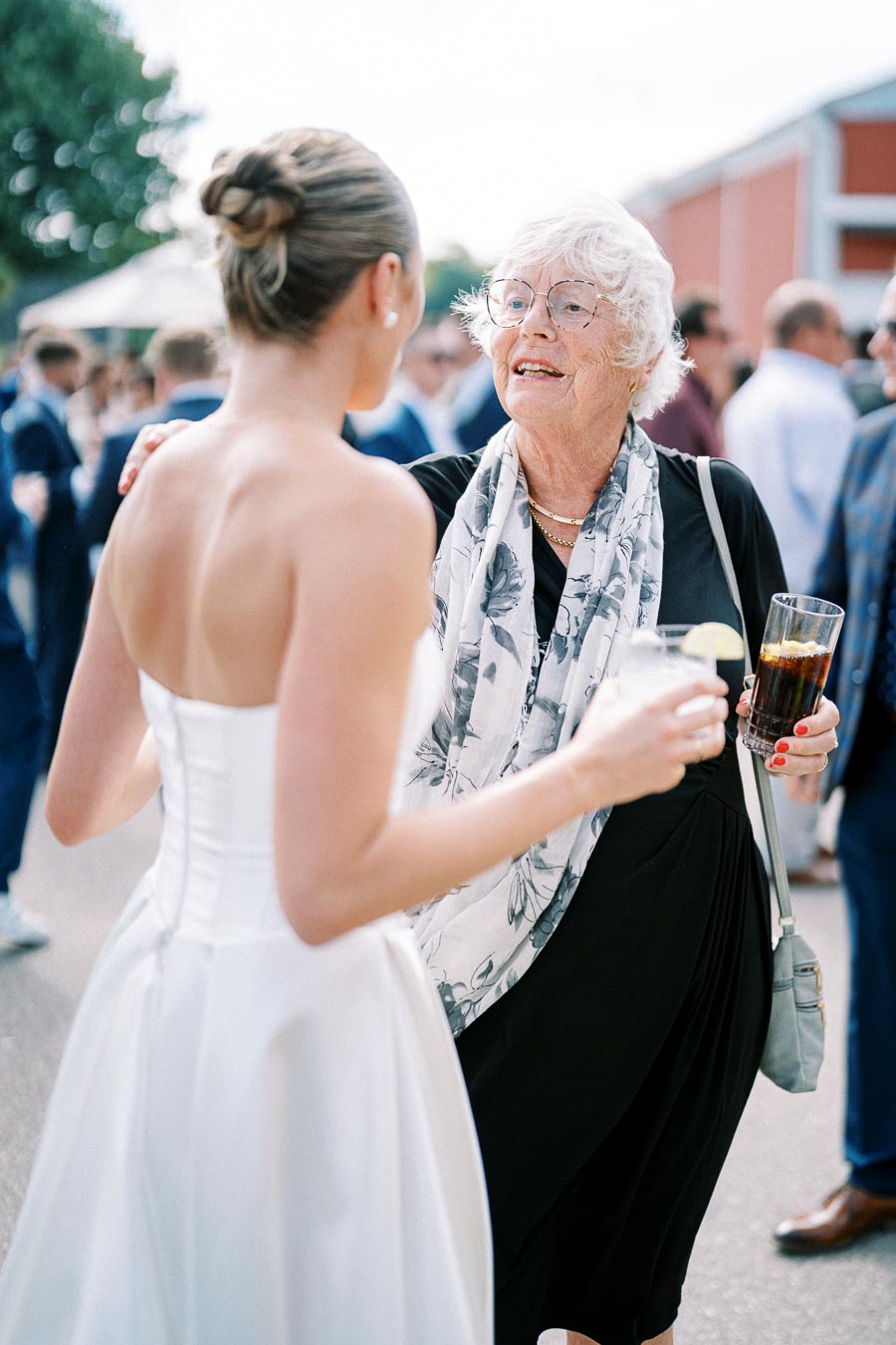 A young woman in a white dress interacts with an elderly woman wearing a floral scarf at an outdoor event. Both hold drinks, and people are visible in the background, suggesting a social gathering or celebration.