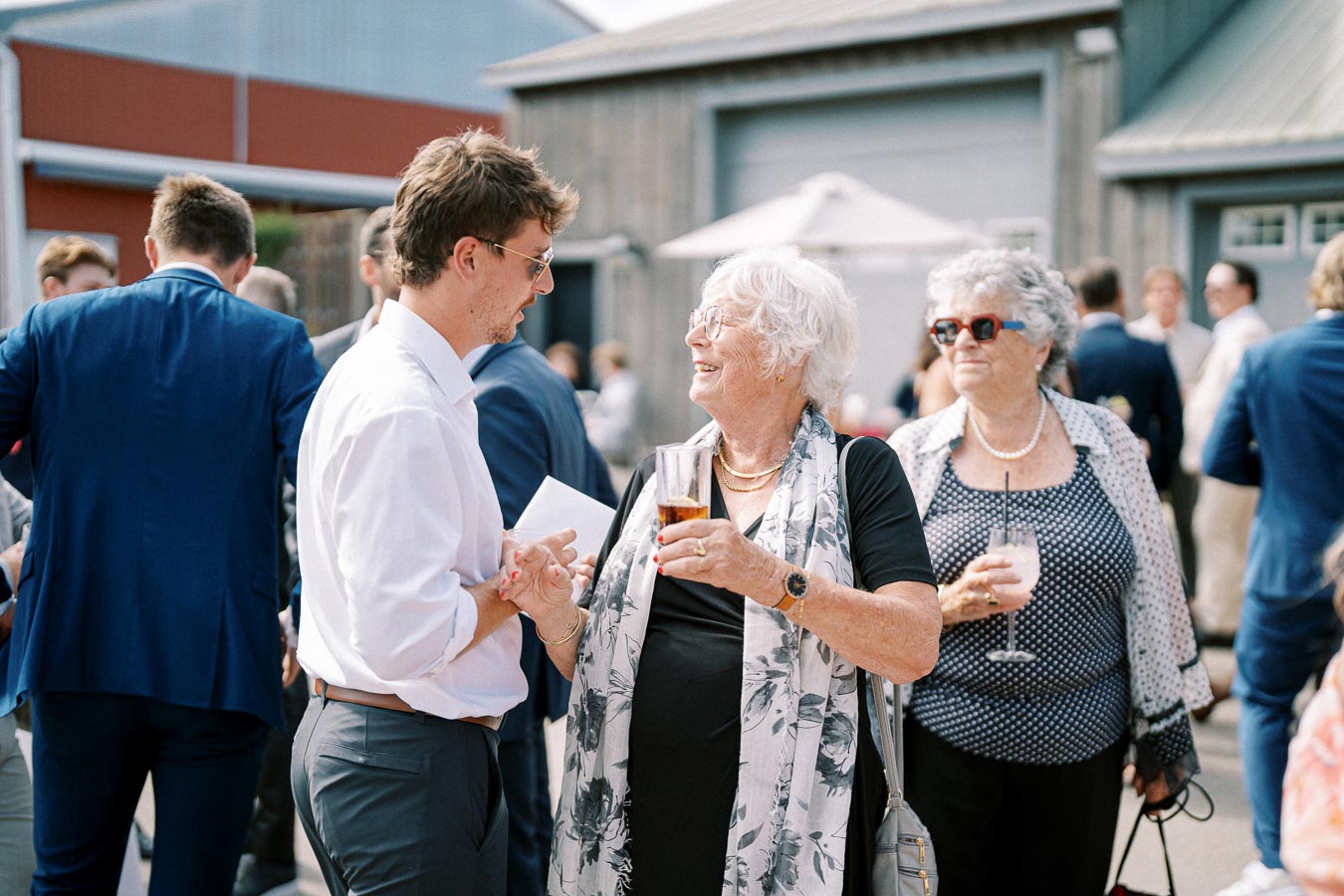 A group of people enjoying a social outdoor gathering, with a woman in glasses holding a drink and engaging in conversation with a man in a white shirt, surrounded by other well-dressed individuals.