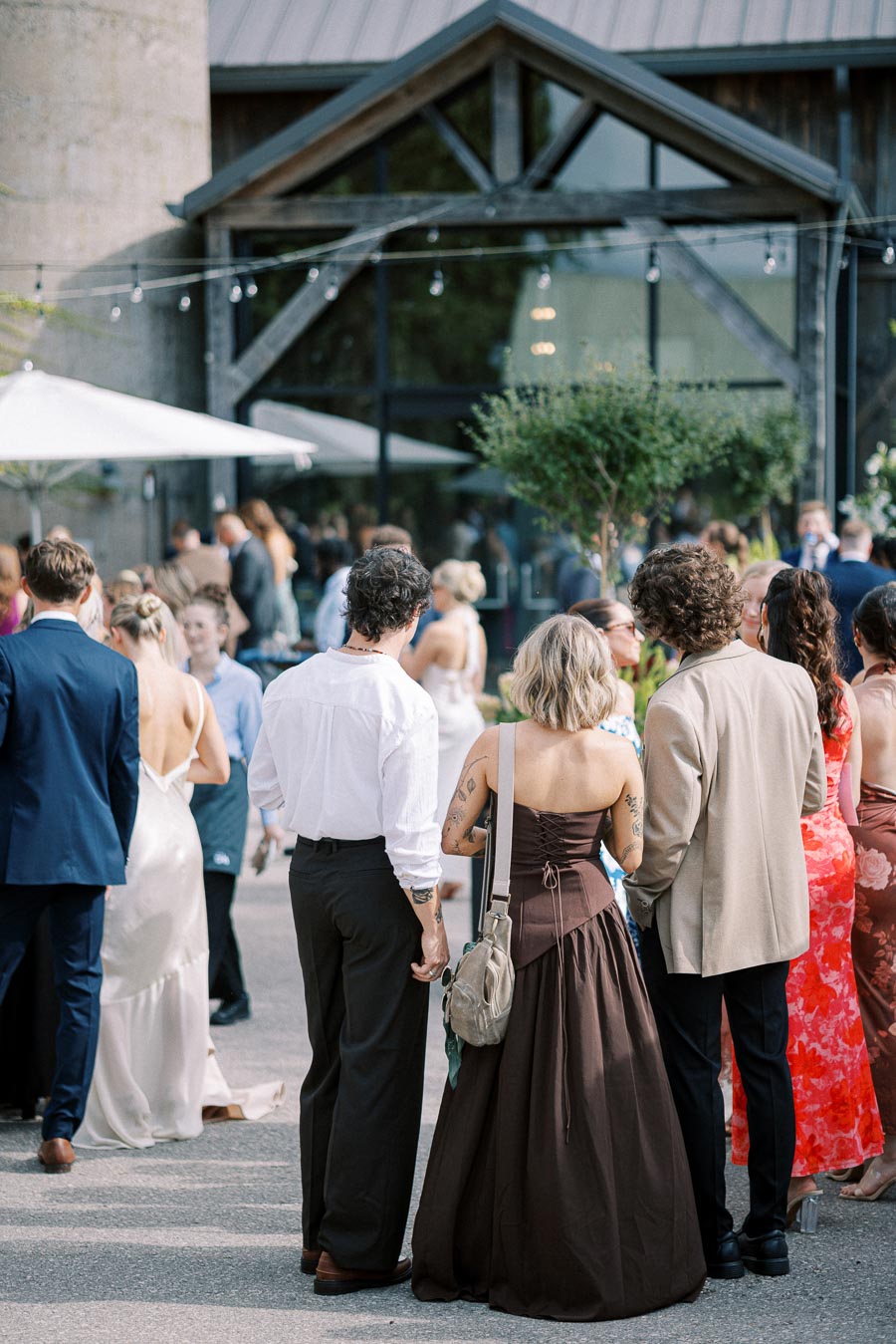 Outdoor wedding reception with guests in formal attire, featuring elegant dresses and suits, gathered near a rustic venue with string lights and greenery.