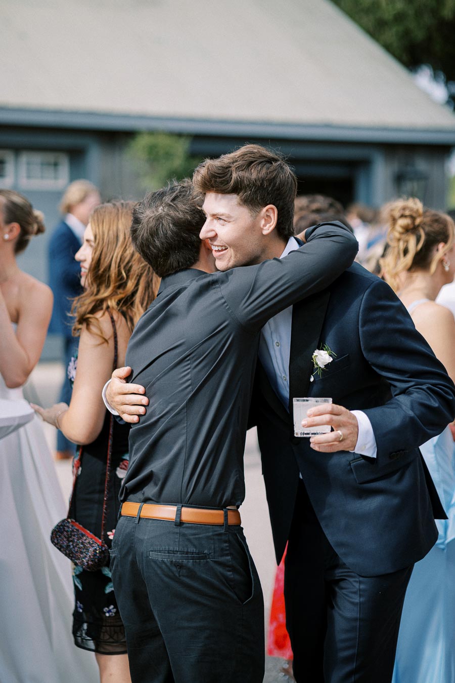 Groom in a navy suit warmly embracing a friend at an outdoor wedding reception, surrounded by smiling guests.