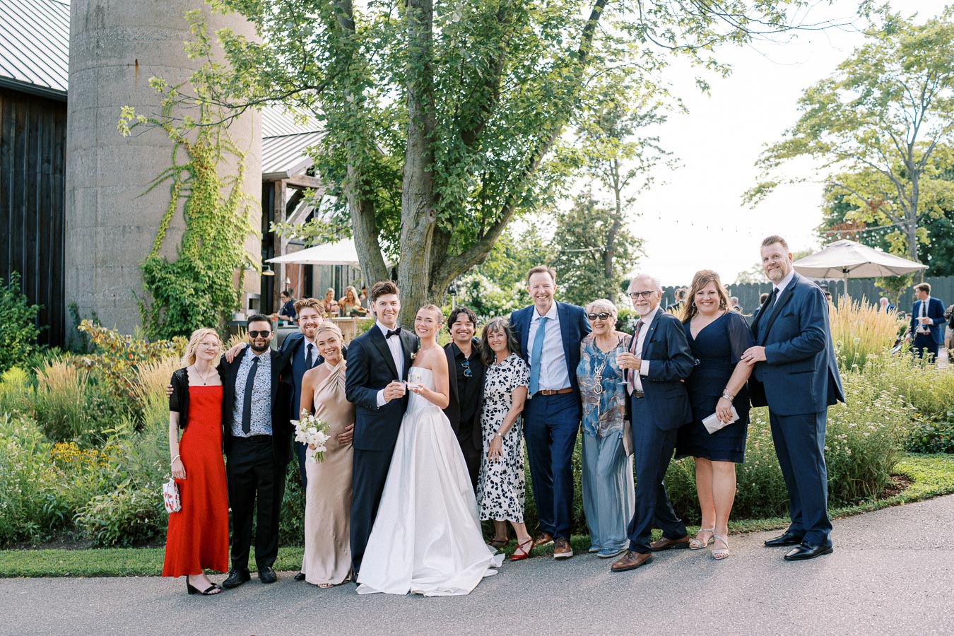 Wedding celebration group photo featuring the bride and groom with family and friends in elegant attire, set in a garden venue with lush greenery and a rustic building backdrop.