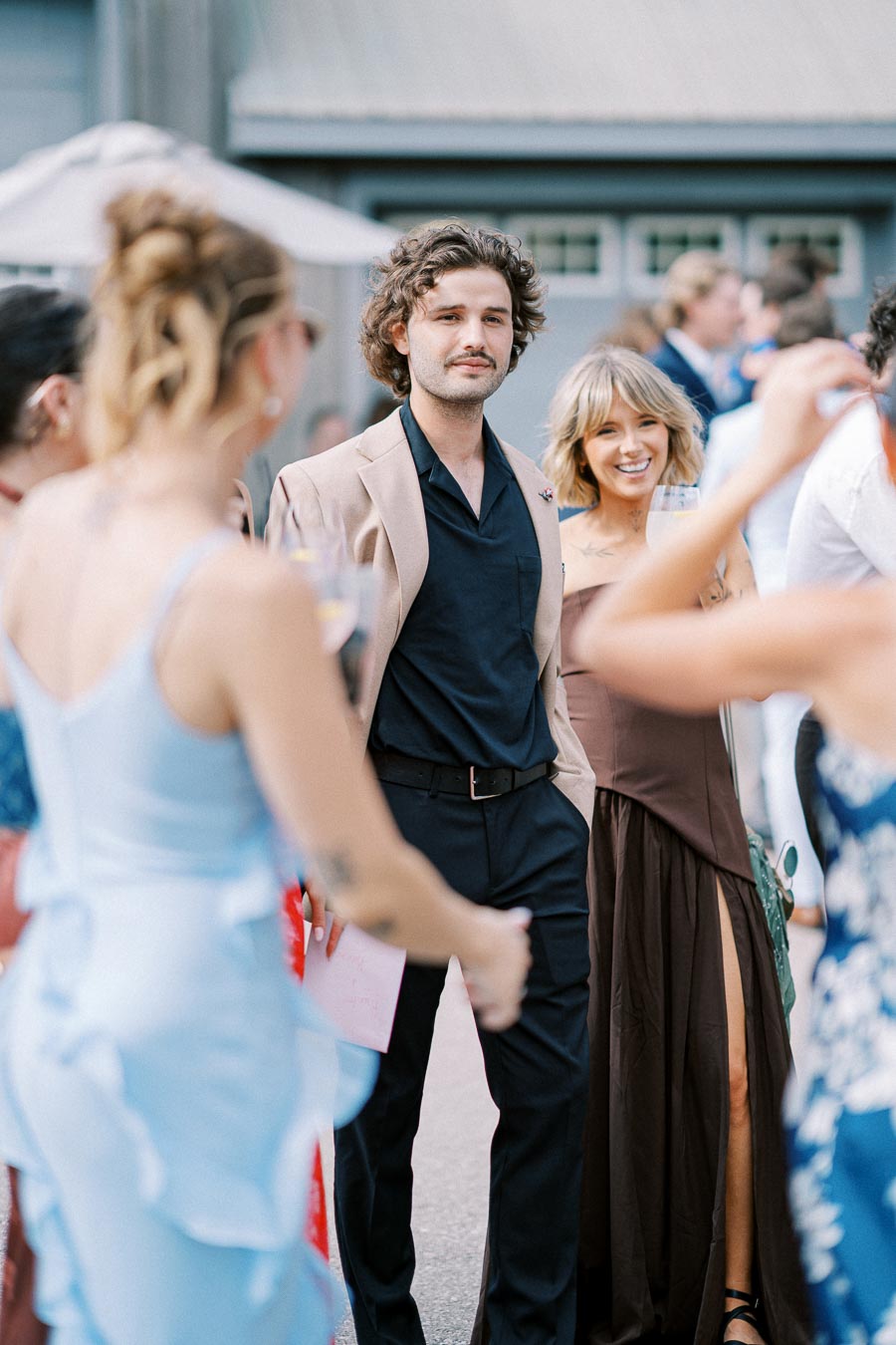 A group of elegantly dressed people socializing at an outdoor event, with a man in a beige jacket and a woman in a brown dress in focus, surrounded by others in vibrant attire.