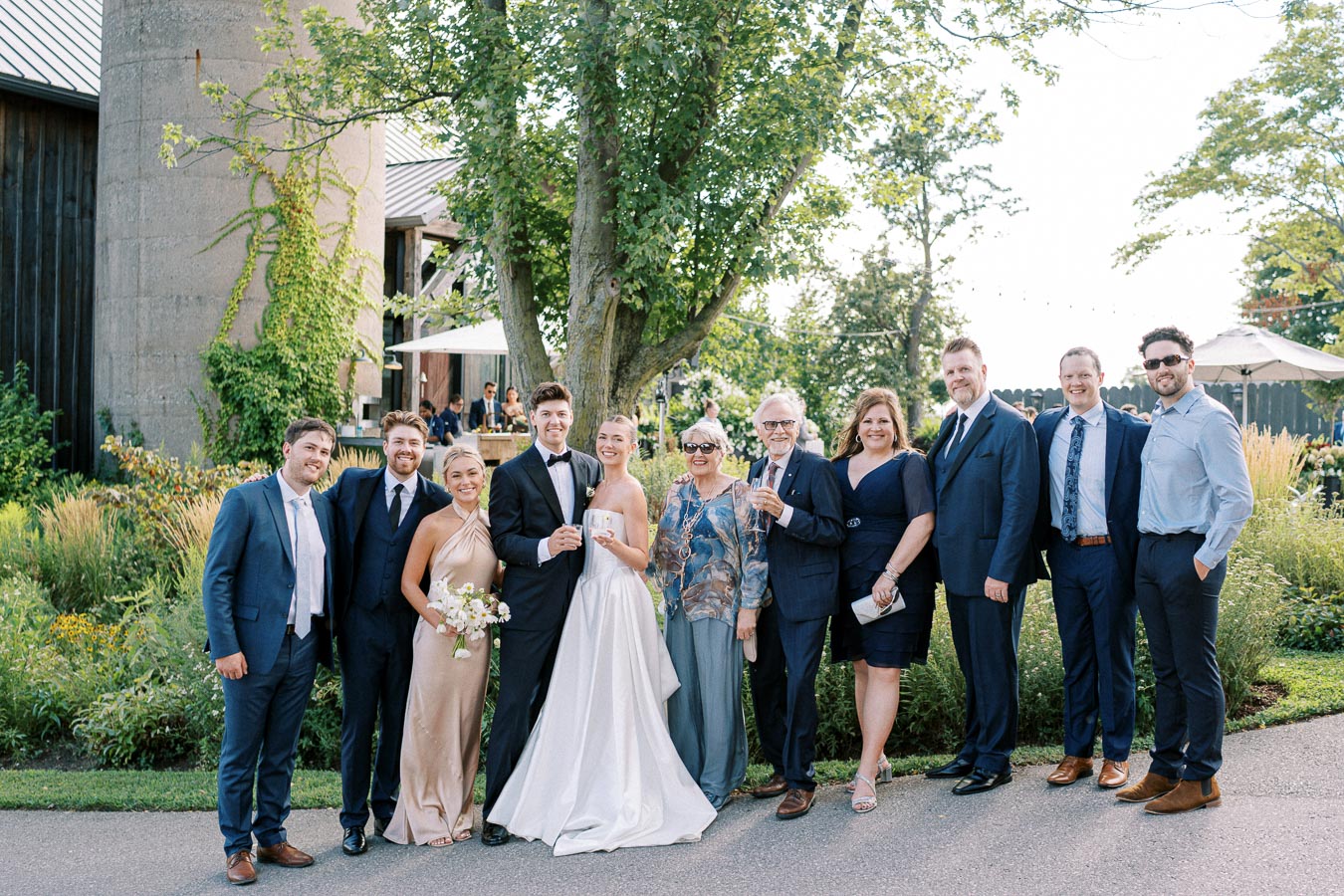 A joyful wedding party poses outdoors, featuring the bride in a white gown and groom in a tuxedo surrounded by elegantly dressed family and friends against a lush garden backdrop.