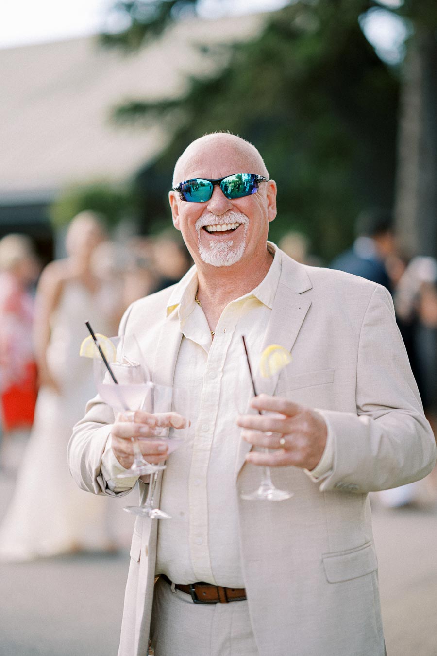 A smiling man in a beige suit and sunglasses holding two cocktail glasses with lemon slices outdoors.