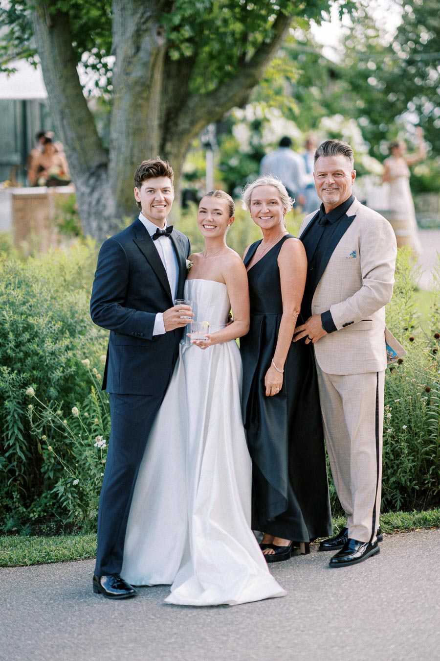 A group of four elegantly dressed people smiling outdoors at a wedding. The woman in a white bridal gown stands beside a man in a black tuxedo, while another woman in a black dress and a man in a beige suit join them. The lush green background suggests a garden setting.