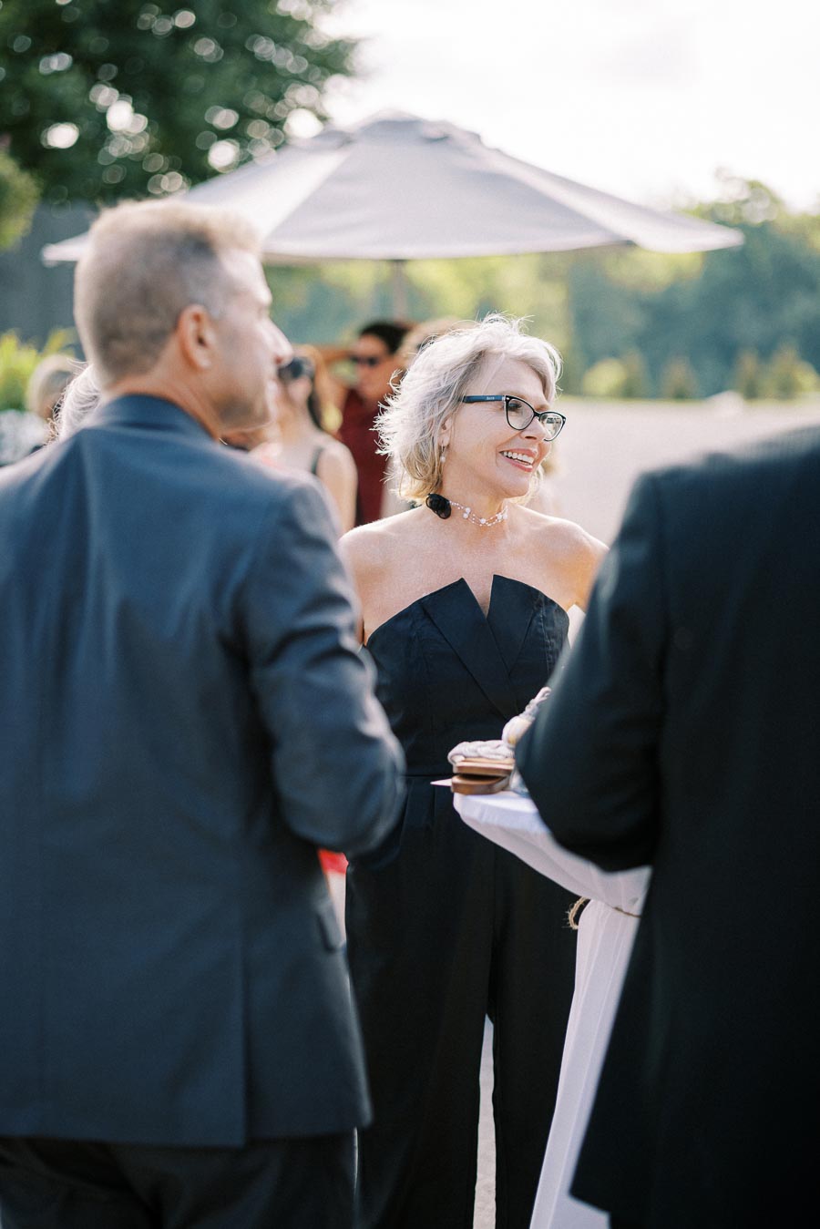 Elegant outdoor social gathering with a smiling woman in a black dress and glasses, conversing with others under a large umbrella in a sunny setting.