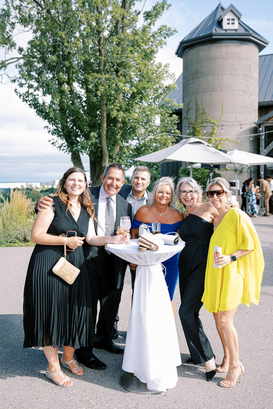 A group of six people dressed in formal and semi-formal attire enjoying an outdoor gathering at a winery, standing near a cocktail table with drinks and smiling, with scenic greenery and a rustic building in the background under a clear sky.
