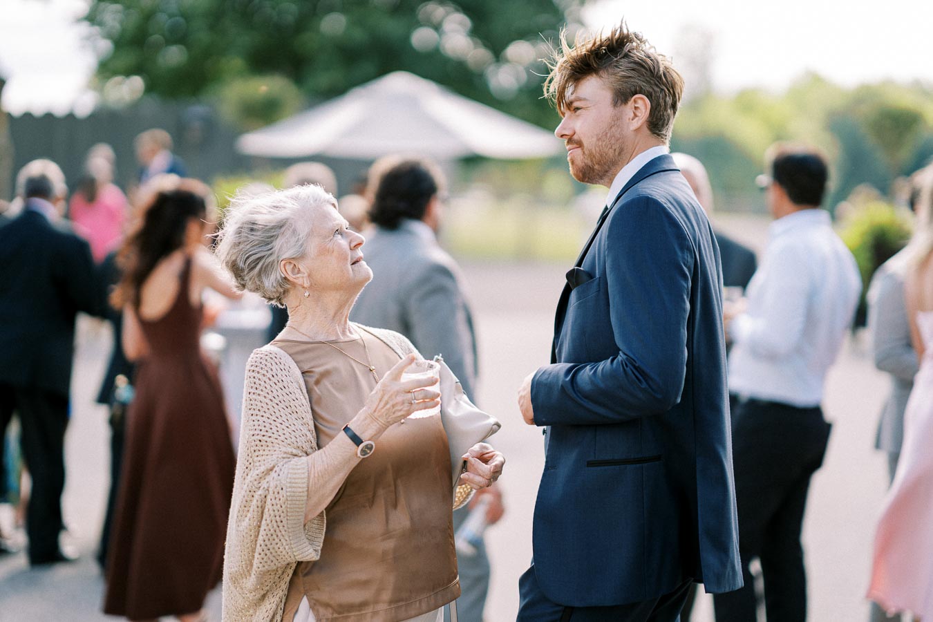 Elderly woman engaging in conversation with a young man in a blue suit at an outdoor social event, with blurred guests in the background.