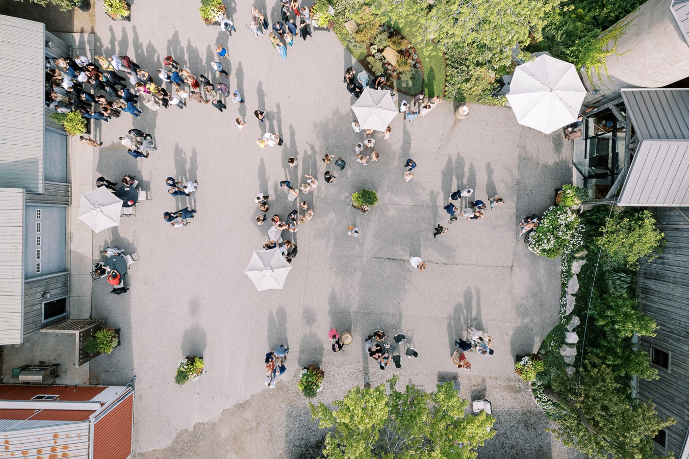 Aerial view of a large group of people gathered in an outdoor courtyard. The scene includes several white umbrellas providing shade, along with greenery and nearby buildings, creating a lively social atmosphere.