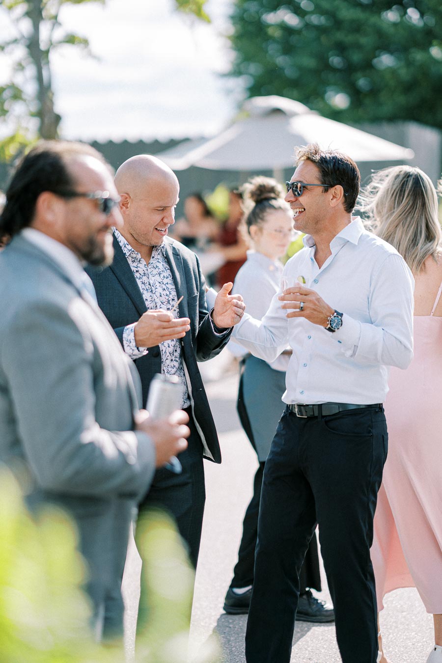 Group of people enjoying a lively outdoor event, dressed in formal attire, socializing and laughing under a sunny sky.