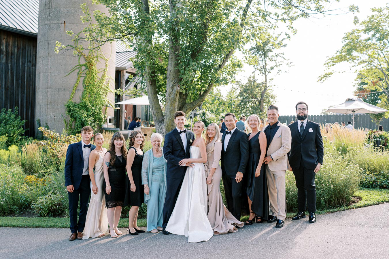 A group of people dressed in formal attire, including a bride and groom, pose together at an outdoor wedding surrounded by greenery and rustic architecture.