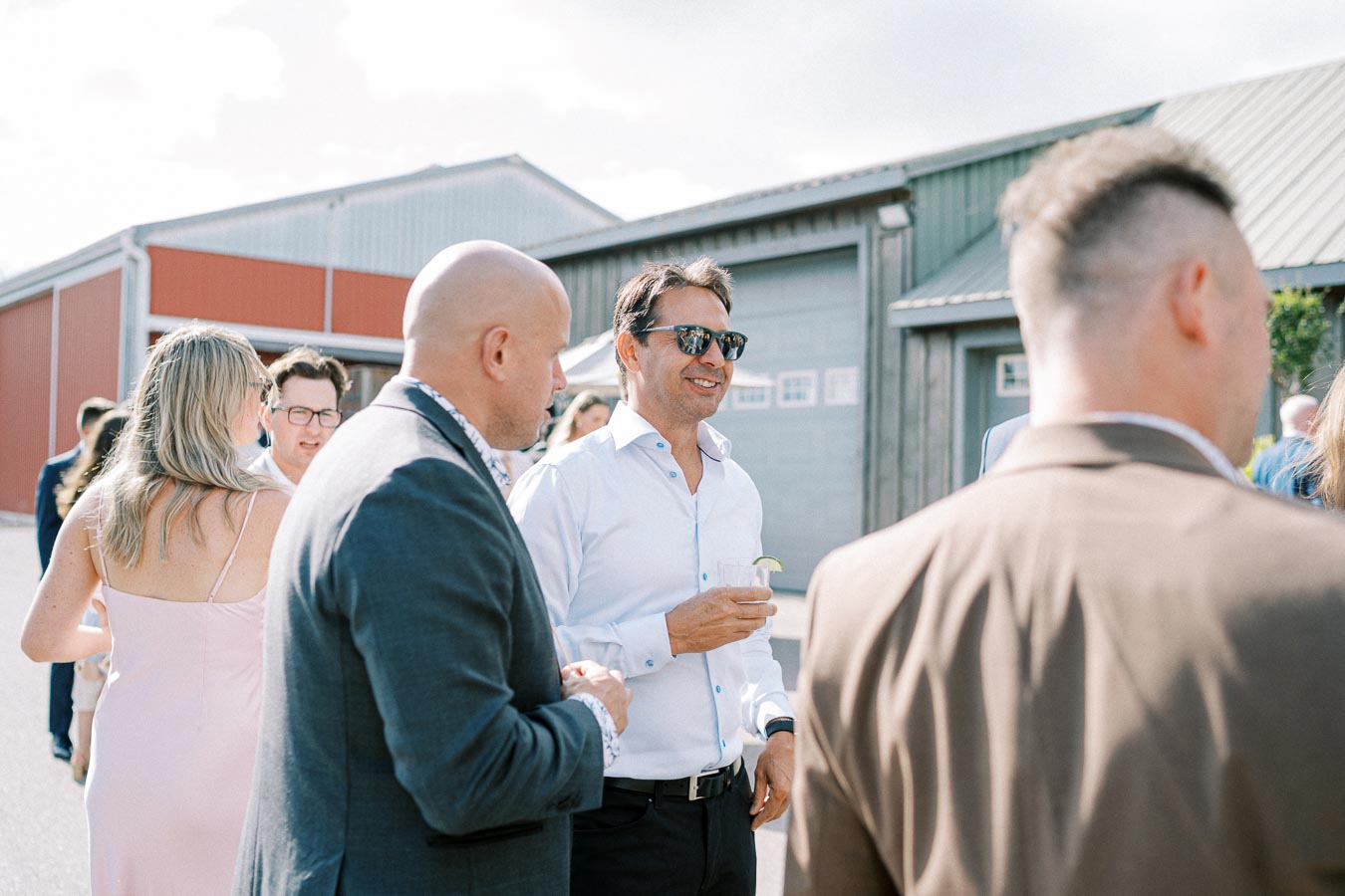 A group of people socializing outdoors at an event, with a man in a white shirt and sunglasses smiling while holding a drink. The setting appears informal, with garage-style buildings in the background under a cloudy sky.