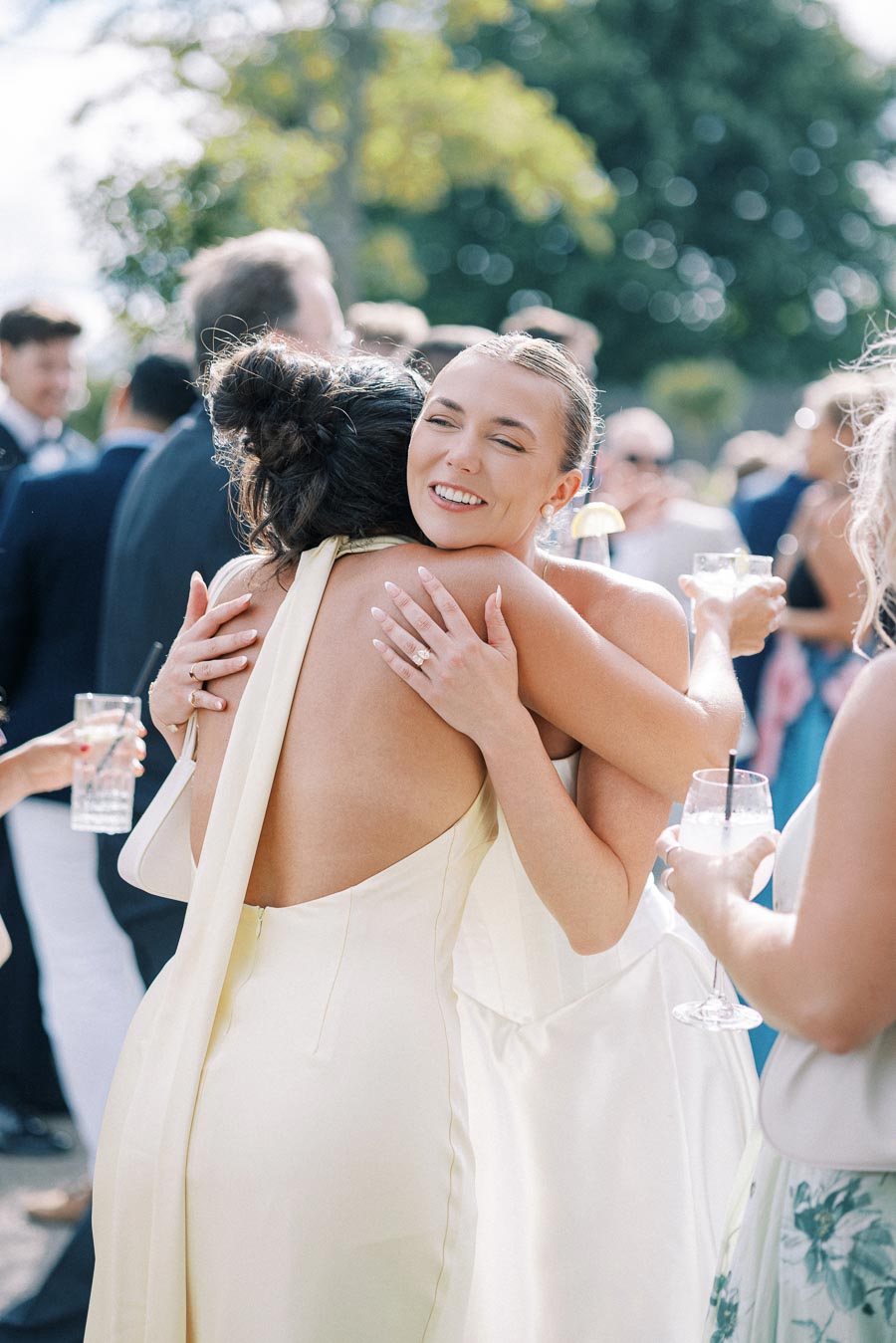Two women in elegant dresses embrace happily at an outdoor wedding reception, surrounded by guests with drinks under sunny skies.