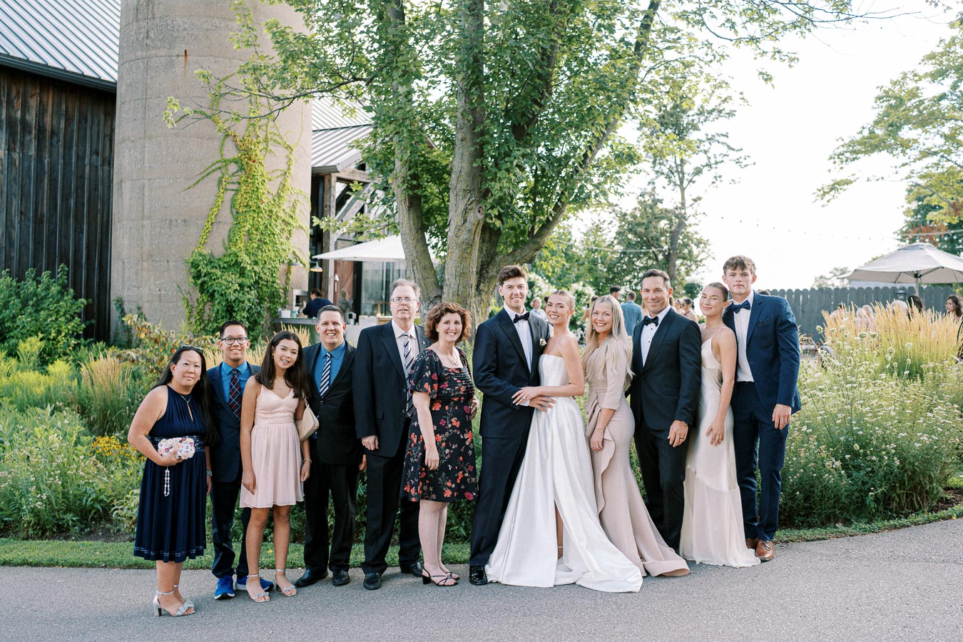A group of people dressed in formal attire stands together in a garden setting, posing for a photo. Elegant outfits and greenery create a celebratory atmosphere, ideal for an outdoor event like a wedding or family gathering.