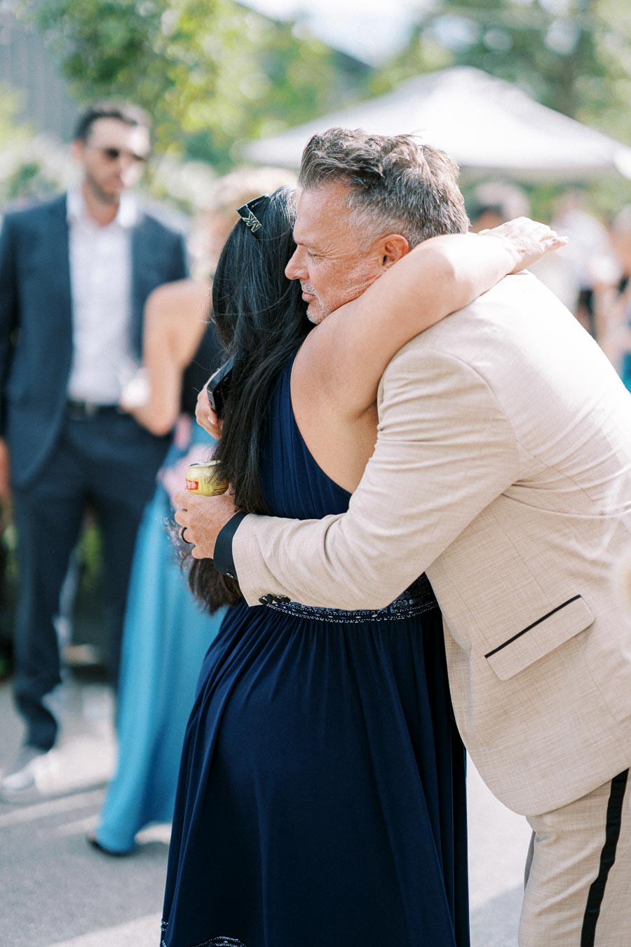 A man in a beige suit embraces a woman in a blue dress at an outdoor event, surrounded by well-dressed guests.