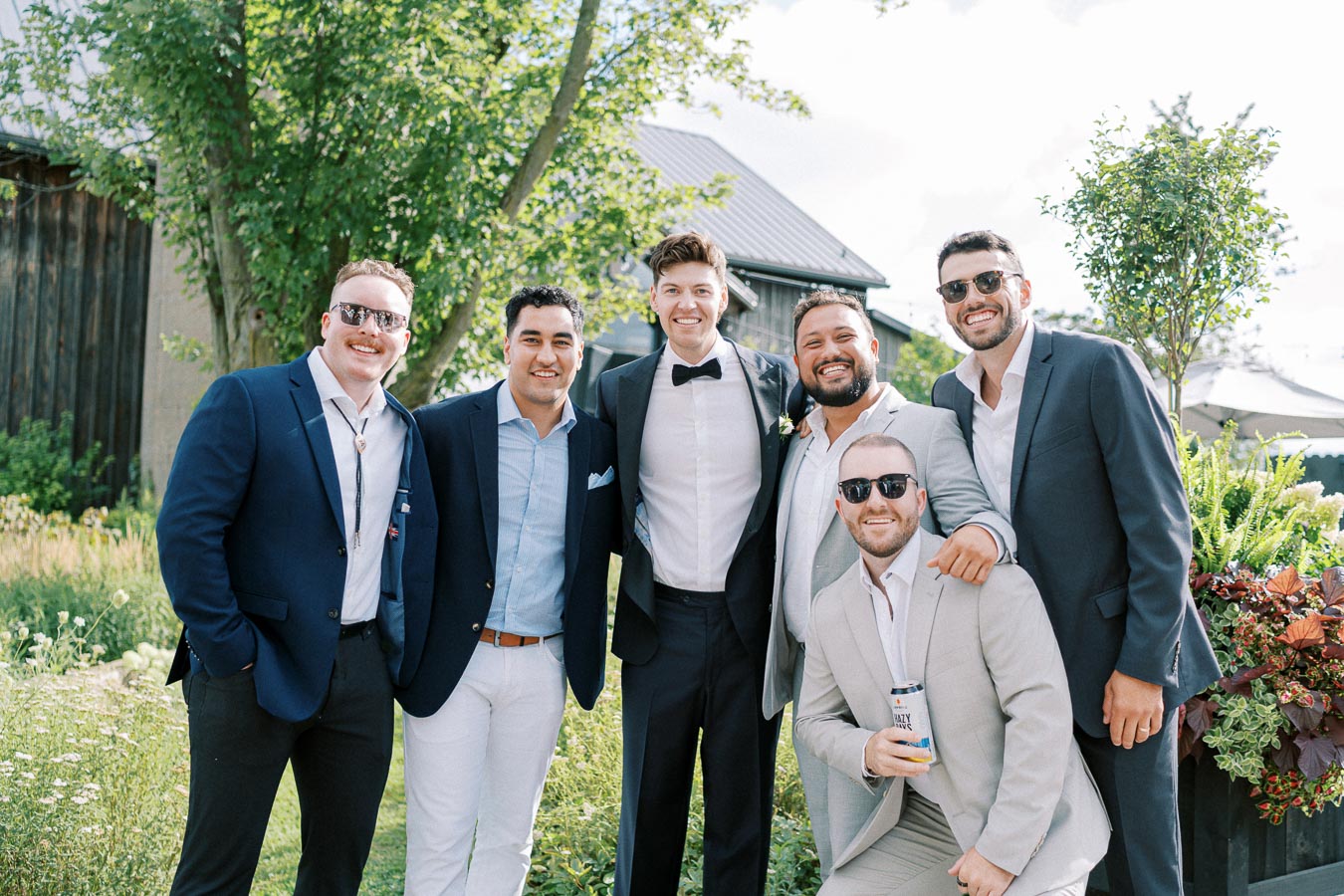 Group of six men in formal attire posing outdoors on a sunny day, surrounded by greenery, with one holding a beverage can.