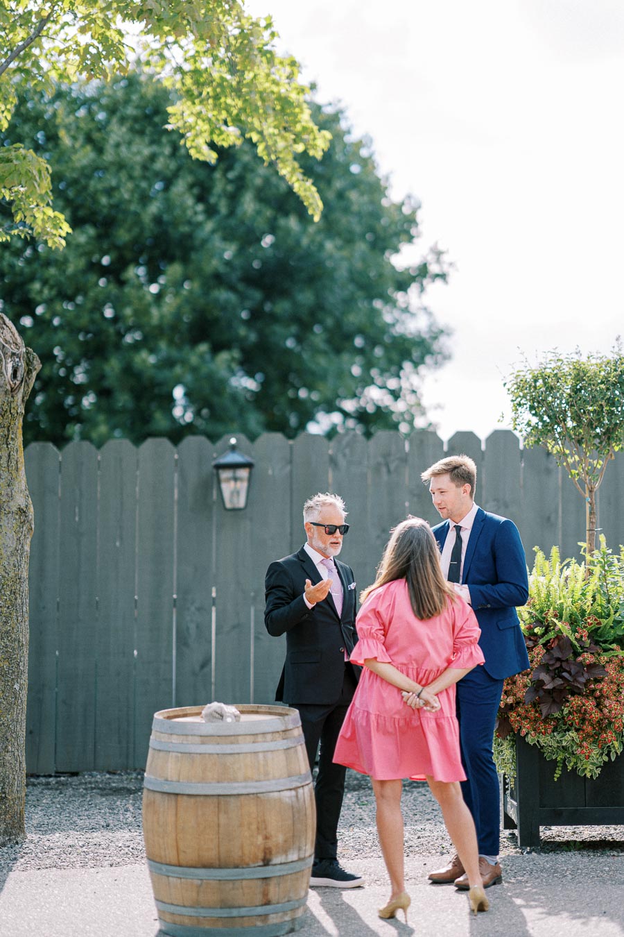 Three people engaged in conversation outdoors, next to a wooden barrel and decorative plants, with a wooden fence and trees in the background. One person wears a pink dress, and the others are in suits.