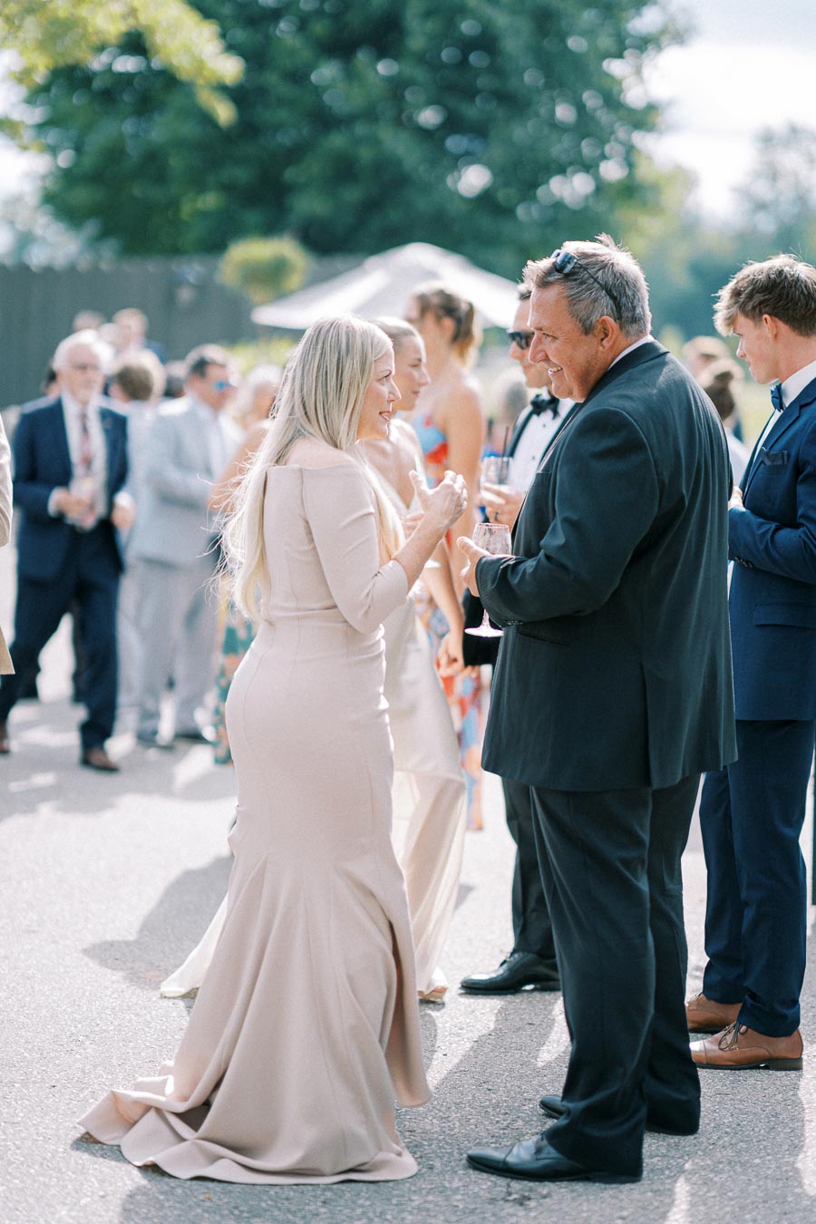 Outdoor wedding reception scene with elegantly dressed guests socializing, featuring a woman in a beige gown and a man in a dark suit holding drinks, set against a sunny backdrop.