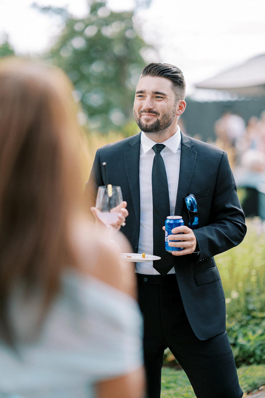 A man in a suit holding a drink at an outdoor social event, smiling and engaging in conversation with a woman in the foreground.