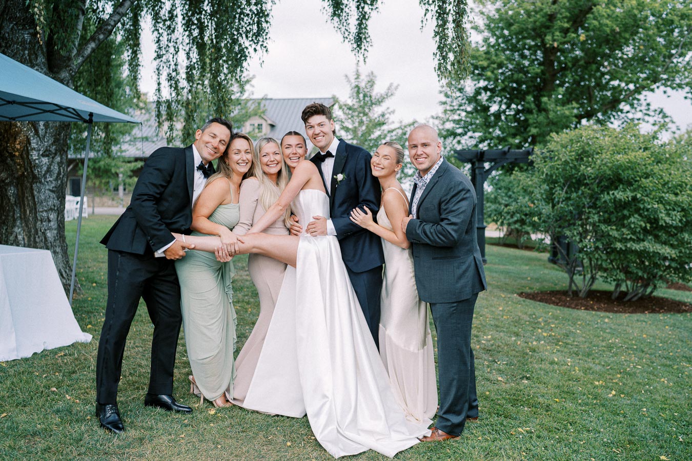 A wedding party joyfully posing in a garden setting, featuring the bride in a white gown held horizontally by the groom and bridesmaids.