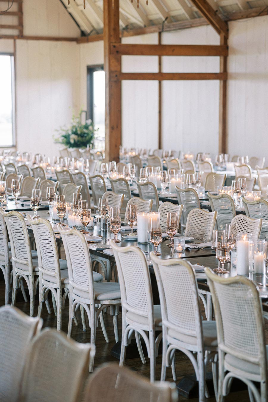 Elegant wedding reception setup in a rustic barn venue, featuring long tables with white chairs, adorned with glassware and candles for a romantic ambiance.