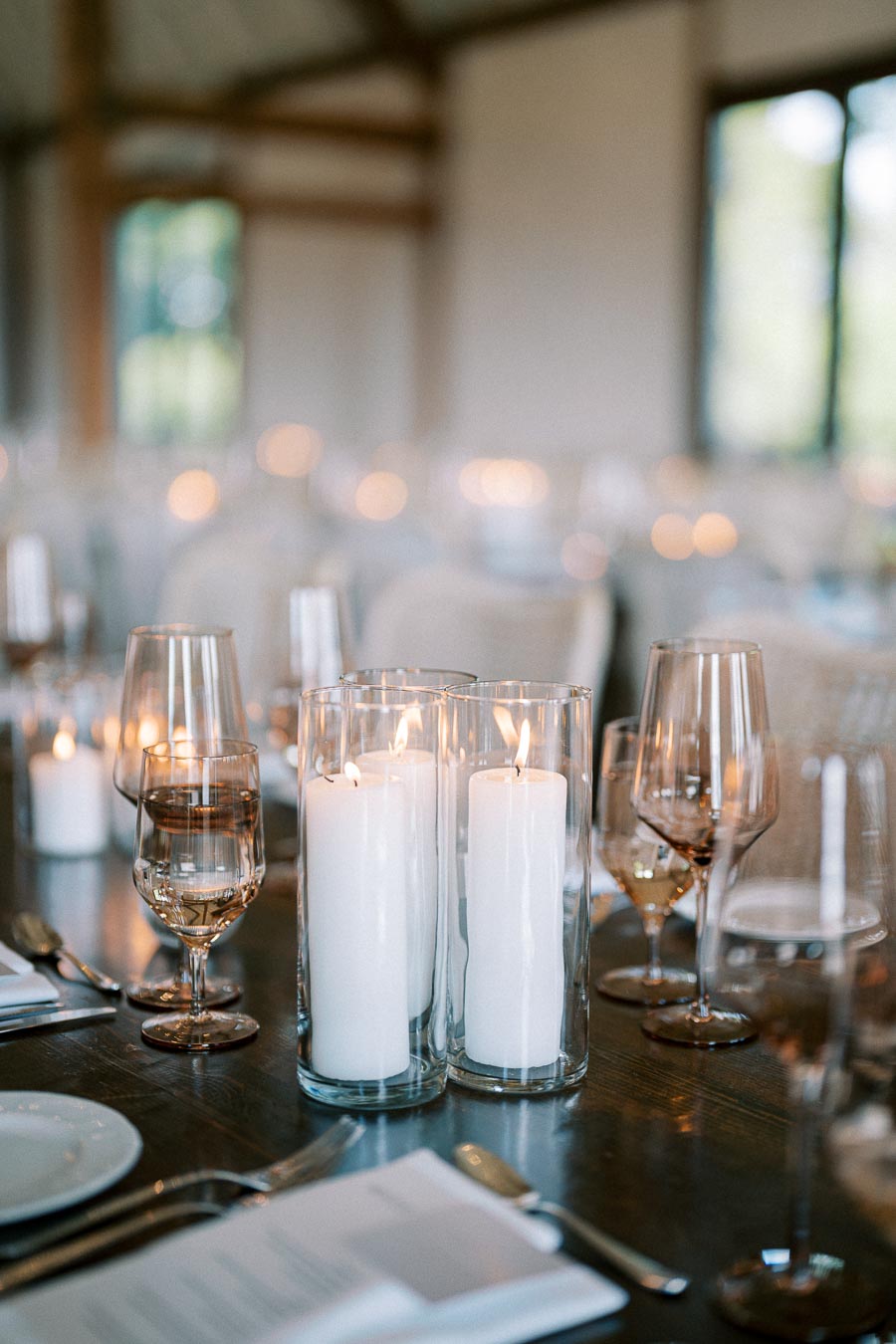 Elegant wedding table setting with tall white candles in glass holders, surrounded by wine glasses and flatware, creating a warm and romantic ambiance.