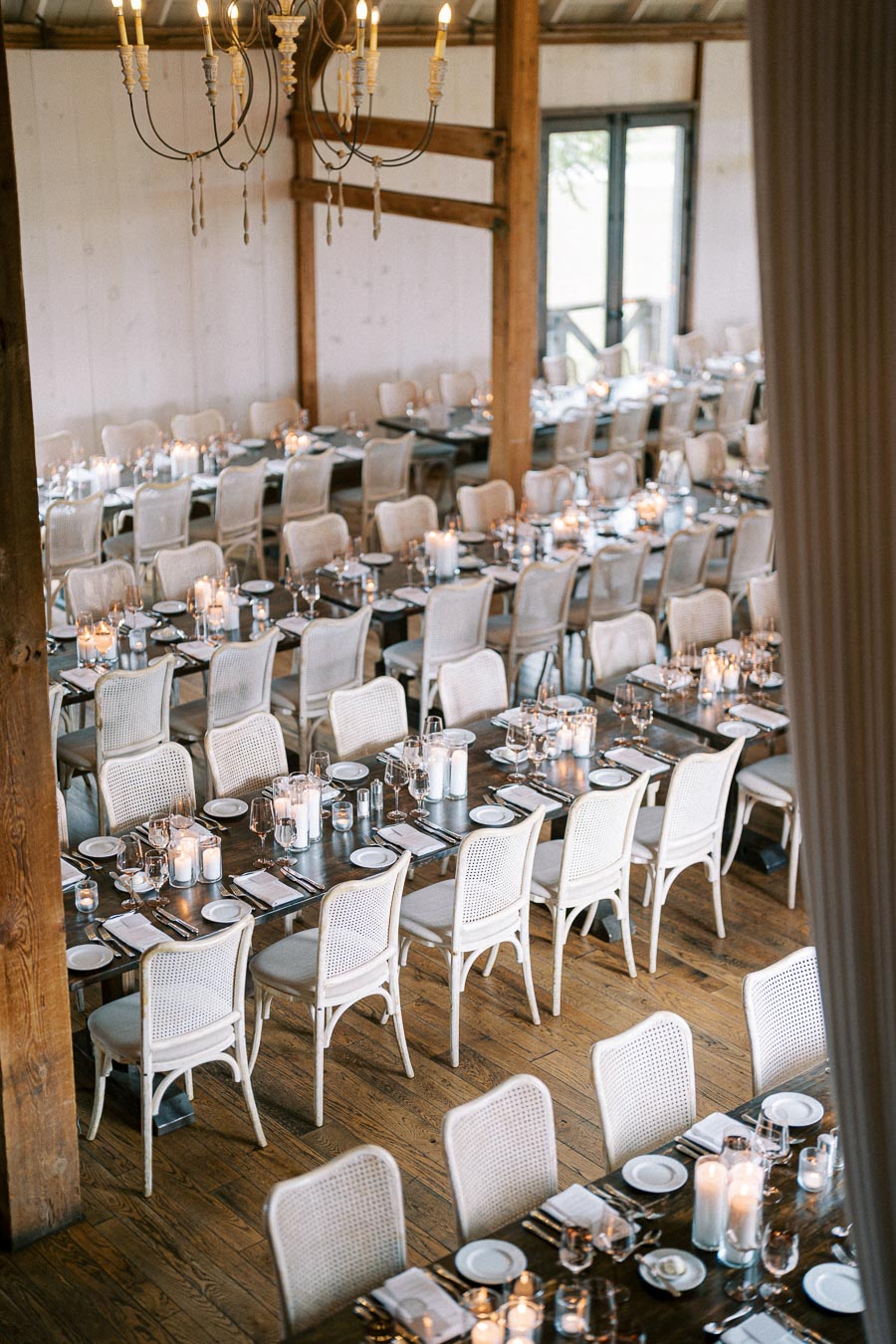 Elegant rustic dining setup in a barn venue, featuring long wooden tables with white chairs arranged for an event. Tables are adorned with candles, glassware, and place settings, creating a warm and inviting atmosphere. A chandelier hangs above, adding a touch of charm and sophistication.
