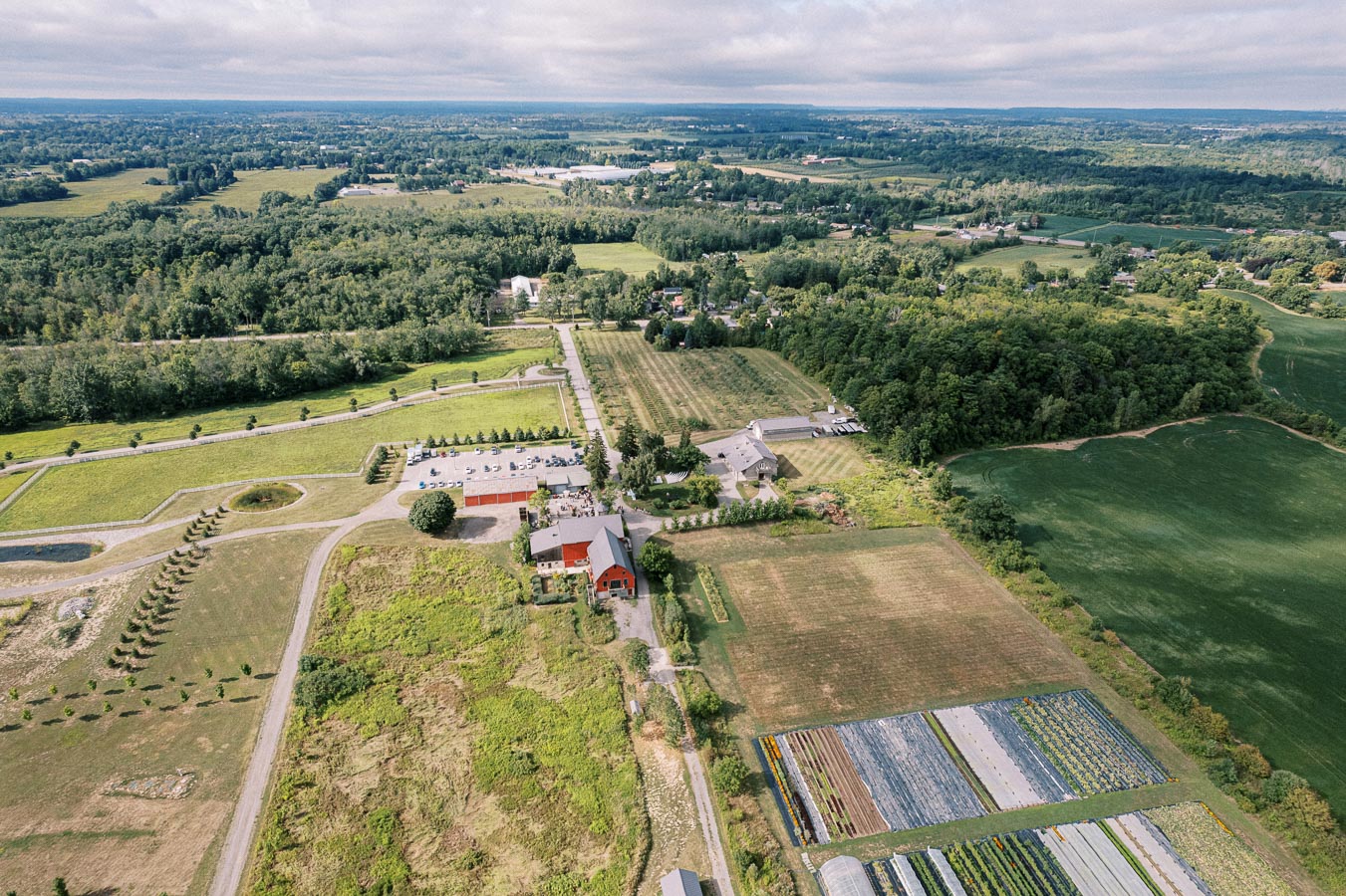 Aerial view of a scenic countryside with sprawling green fields, farm buildings, and winding roads, under a partly cloudy sky.