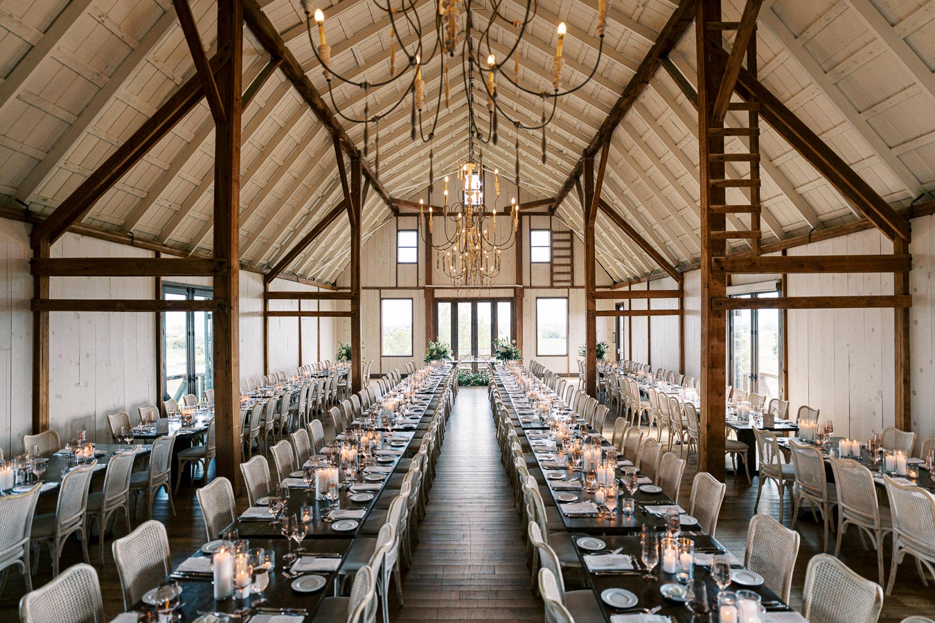 Elegant rustic barn venue with long banquet tables set for a wedding reception, featuring chandeliers, wooden beams, and candlelit ambiance.