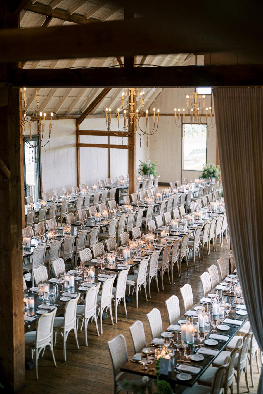 Elegant barn wedding reception setup with long wooden tables, white chairs, and candlelit centerpieces, featuring rustic chandeliers and soft lighting.