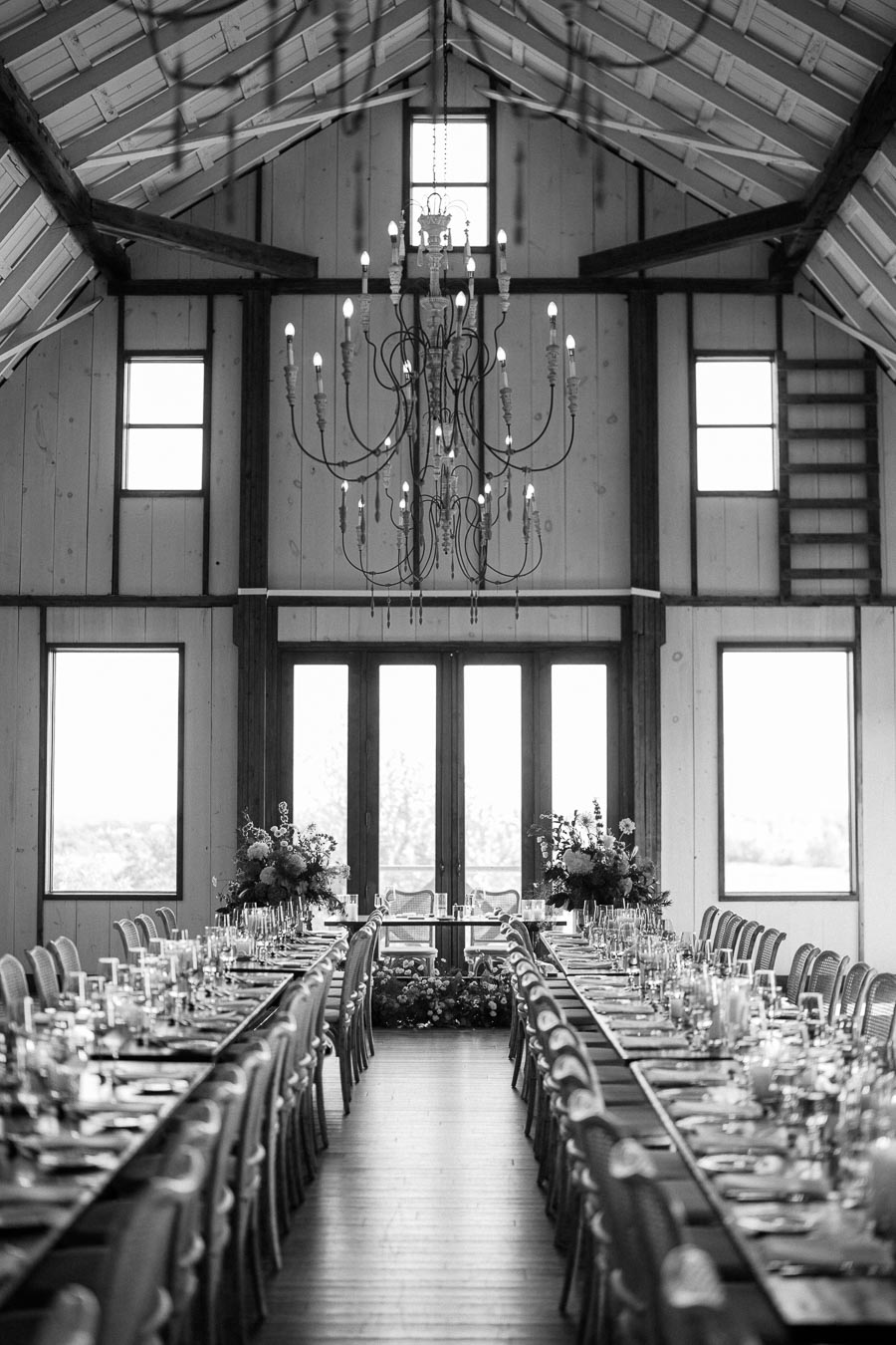 Elegant barn wedding reception setup with a long dining table, chandeliers, and floral arrangements, creating a rustic and romantic atmosphere.
