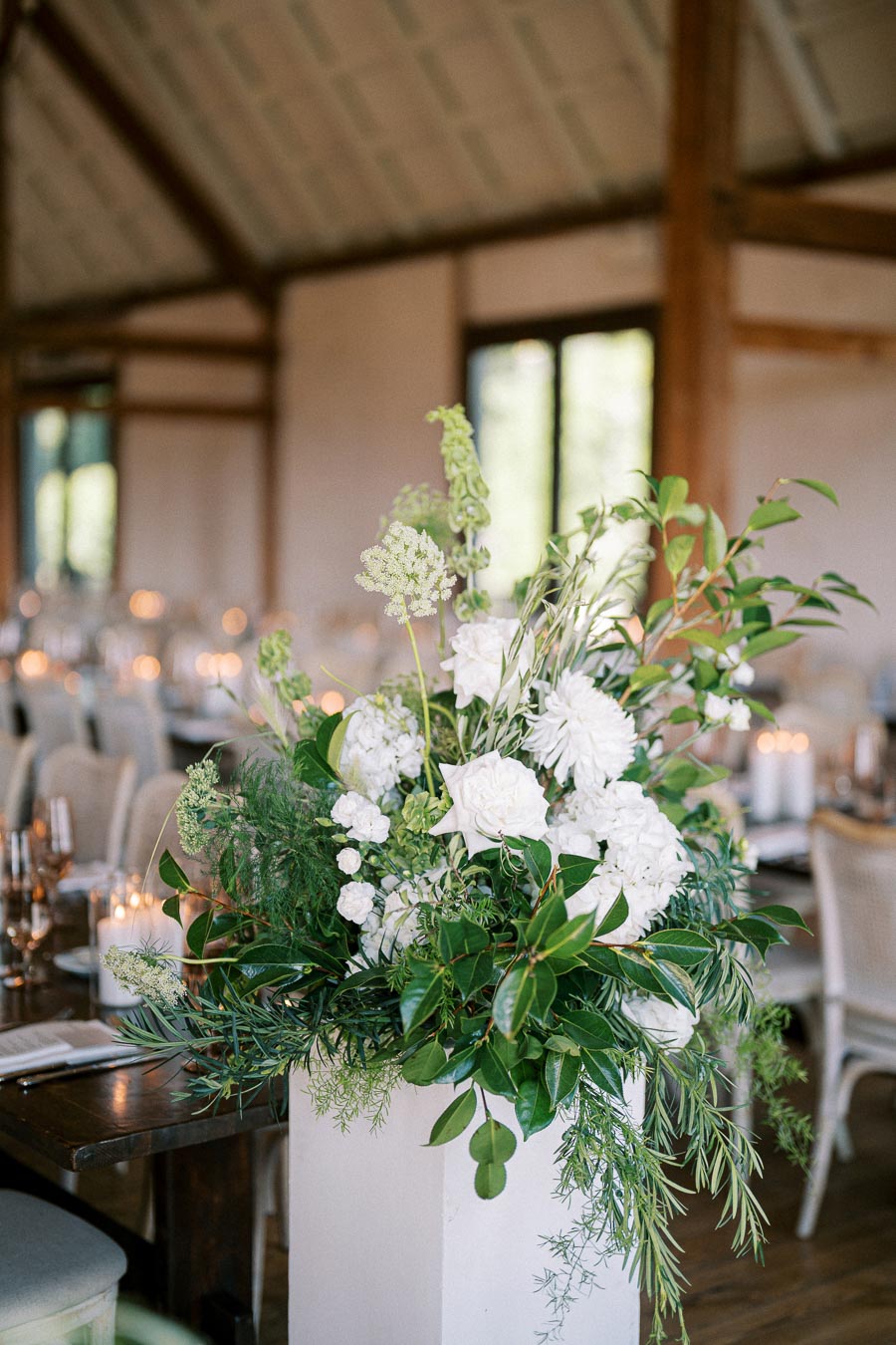 Elegant indoor wedding venue with a large floral arrangement featuring white hydrangeas and greenery, placed on a table with glowing candlelight in the background.