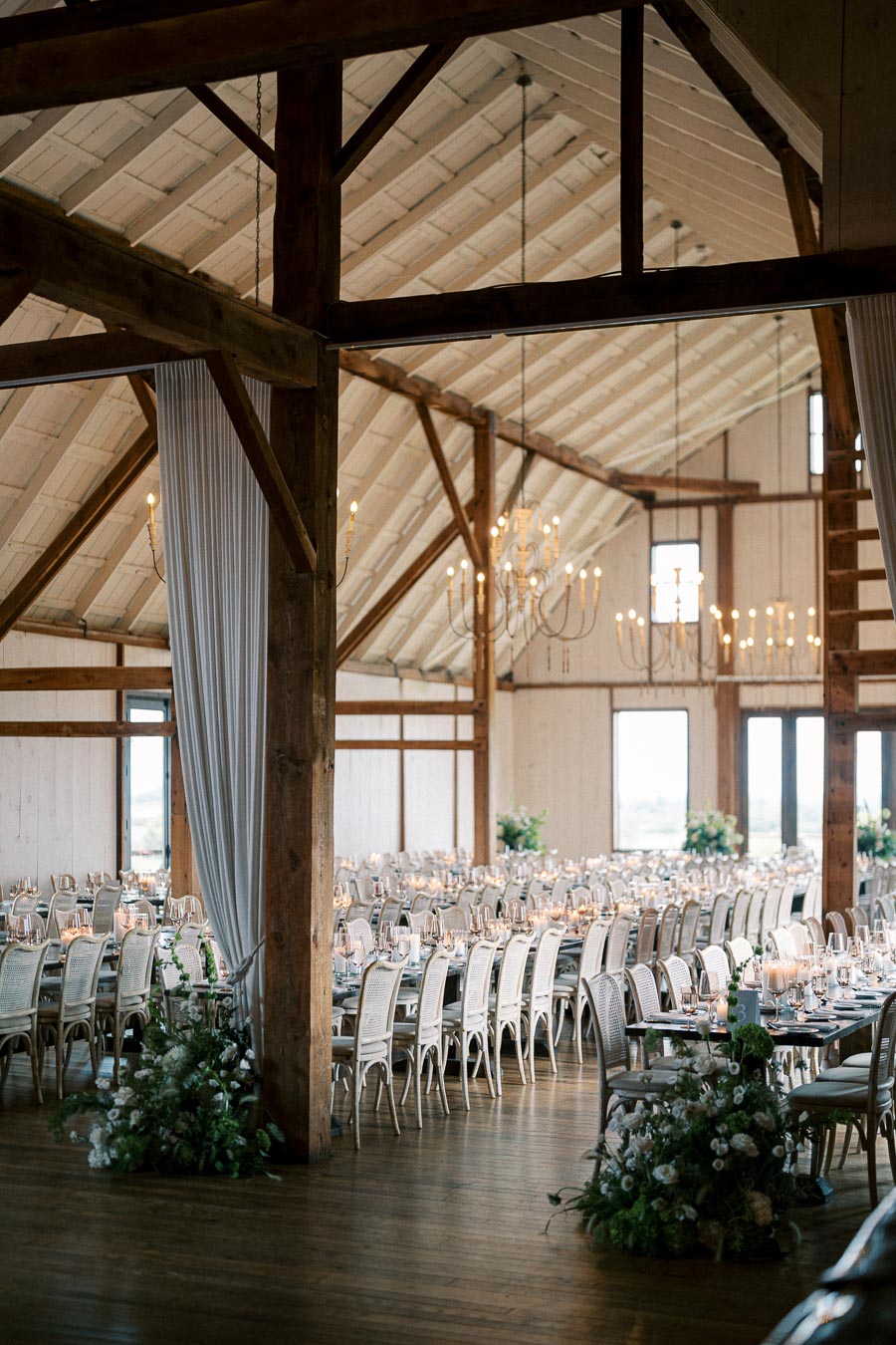 Rustic barn wedding reception venue with elegant chandeliers, white drapery, and long tables adorned with candles and greenery.