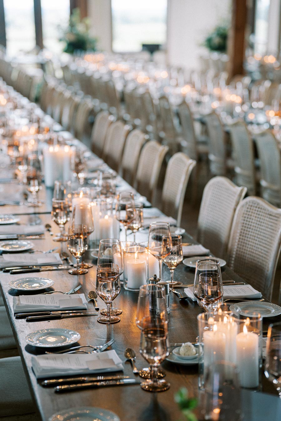 Elegant wedding reception setup with long wooden banquet tables adorned with candles, wine glasses, and delicate tableware in a warmly lit dining hall.