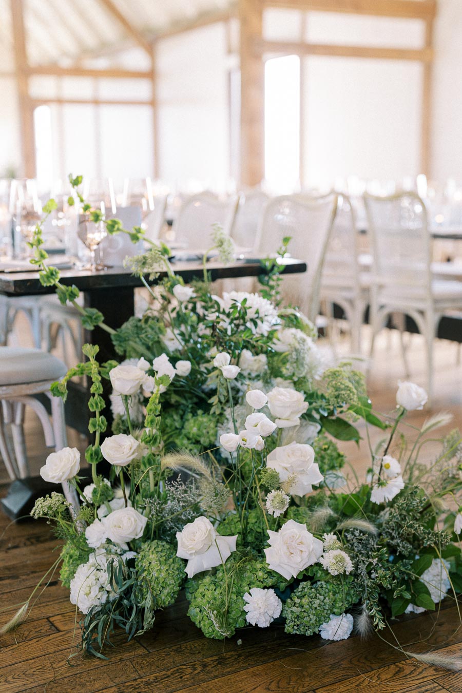 Elegant floral arrangement featuring white roses and greenery decorating a rustic wooden venue with chic white chairs and tables set for a sophisticated event.