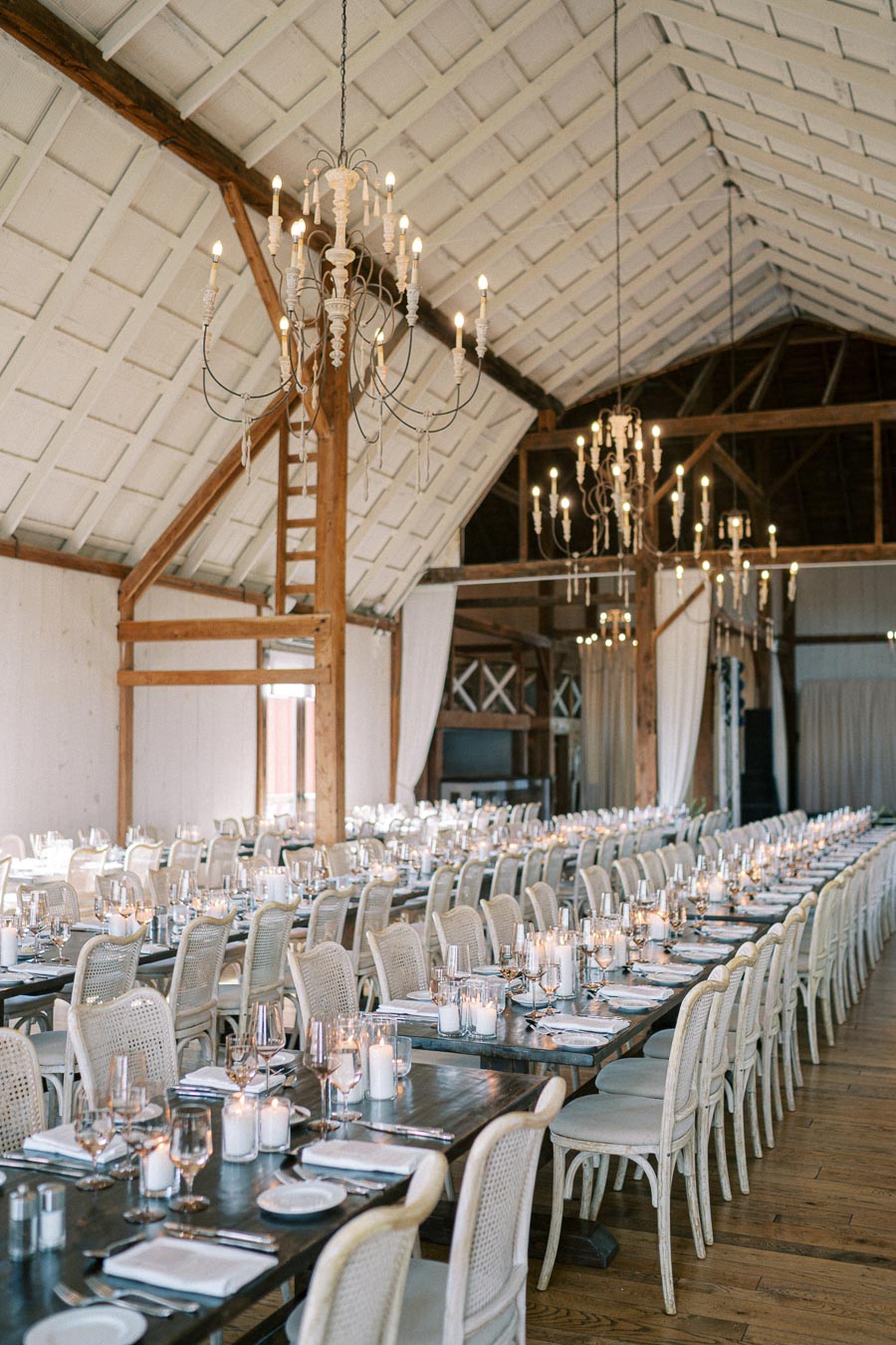 Elegant rustic barn wedding reception setup with long tables, white chairs, and glowing chandeliers.