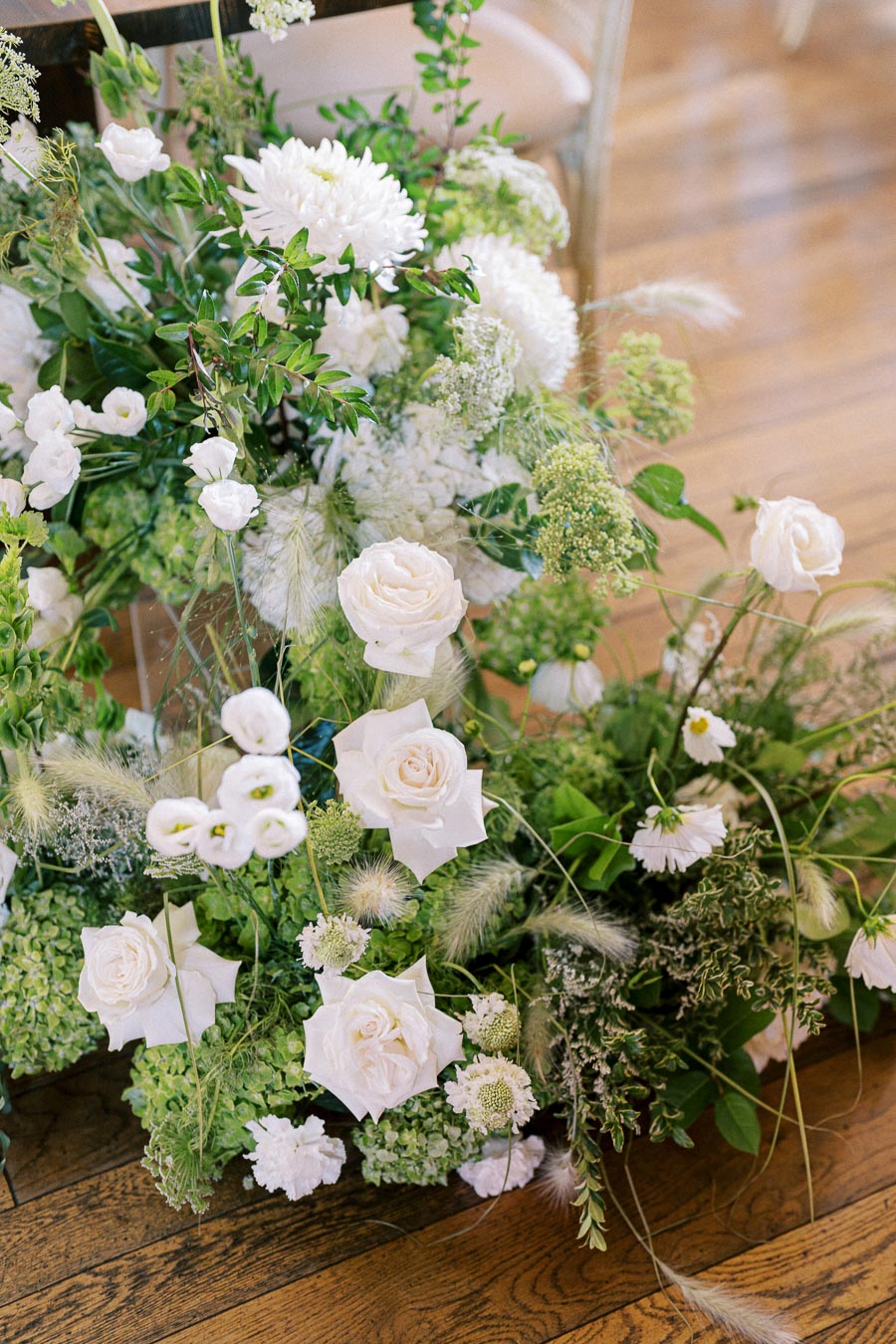 Elegant floral arrangement featuring white roses, lush greenery, and various delicate white flowers, set against a polished wooden floor. Perfect for wedding or event decor inspiration.