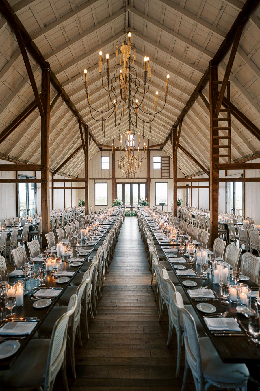 Elegant rustic barn wedding reception setup with long wooden tables, candlelit centerpieces, and hanging chandeliers.