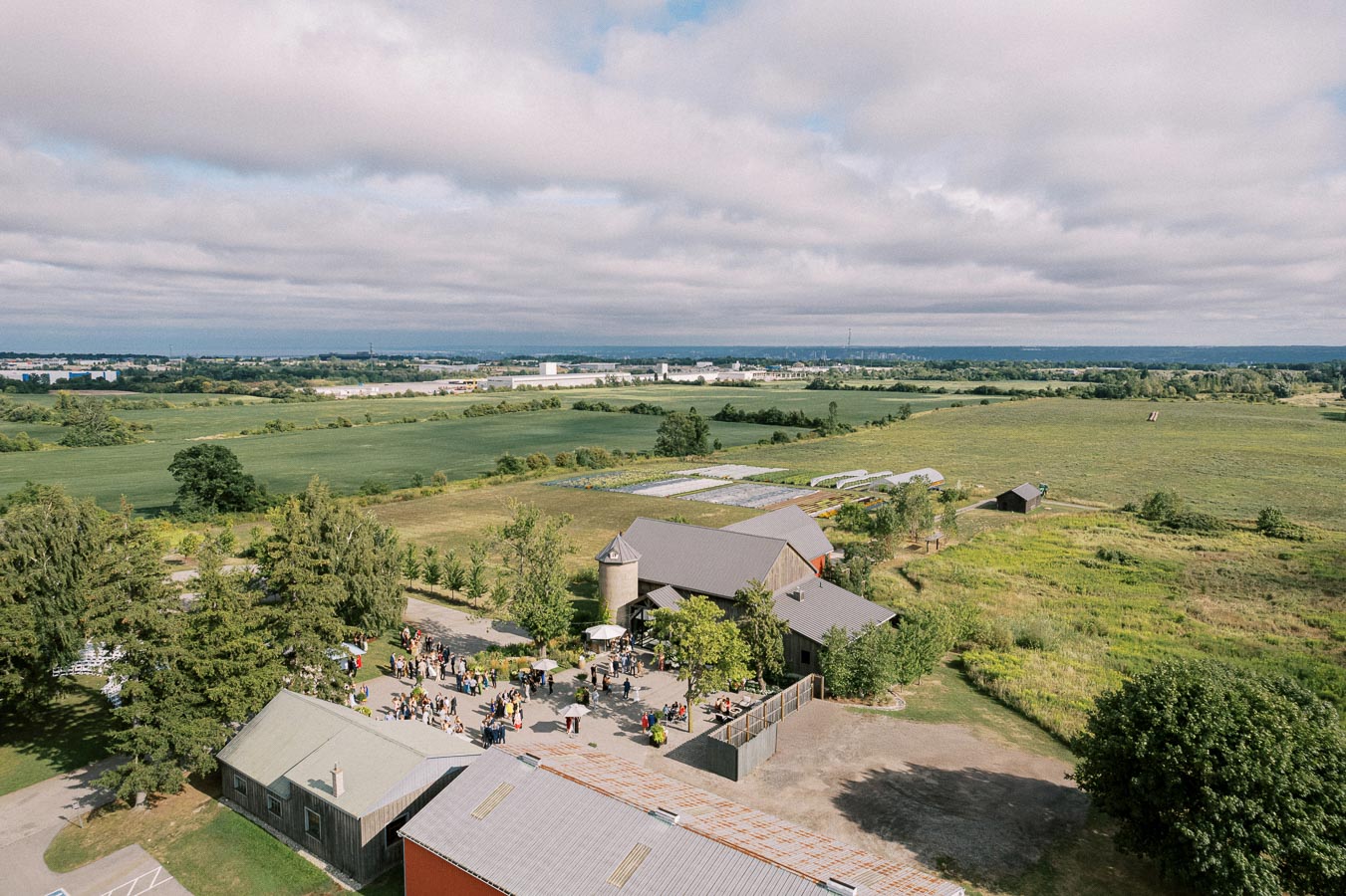 Aerial view of a large farm property with a gathering of people, surrounded by vast green fields under a cloudy sky.