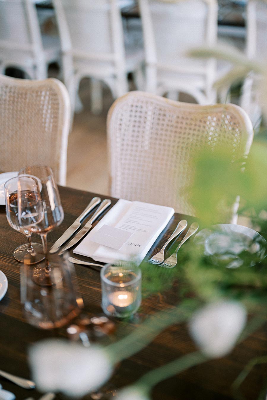 Elegant dining table set for a wedding reception with white cane chairs, crystal glassware, and folded menus on white napkins.