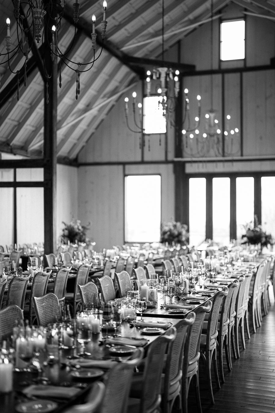 Black and white image of an elegant barn interior set up for a formal event, featuring long wooden tables with decorative place settings and candles, surrounded by stylish chairs, beneath rustic chandeliers.
