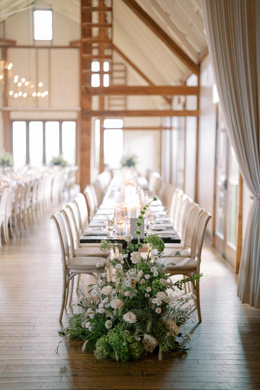 Elegant wedding reception setup in a rustic barn with a long dining table, white chairs, and soft lighting. The table is adorned with candles and lush floral arrangements featuring white and green flowers, creating a romantic and inviting atmosphere.