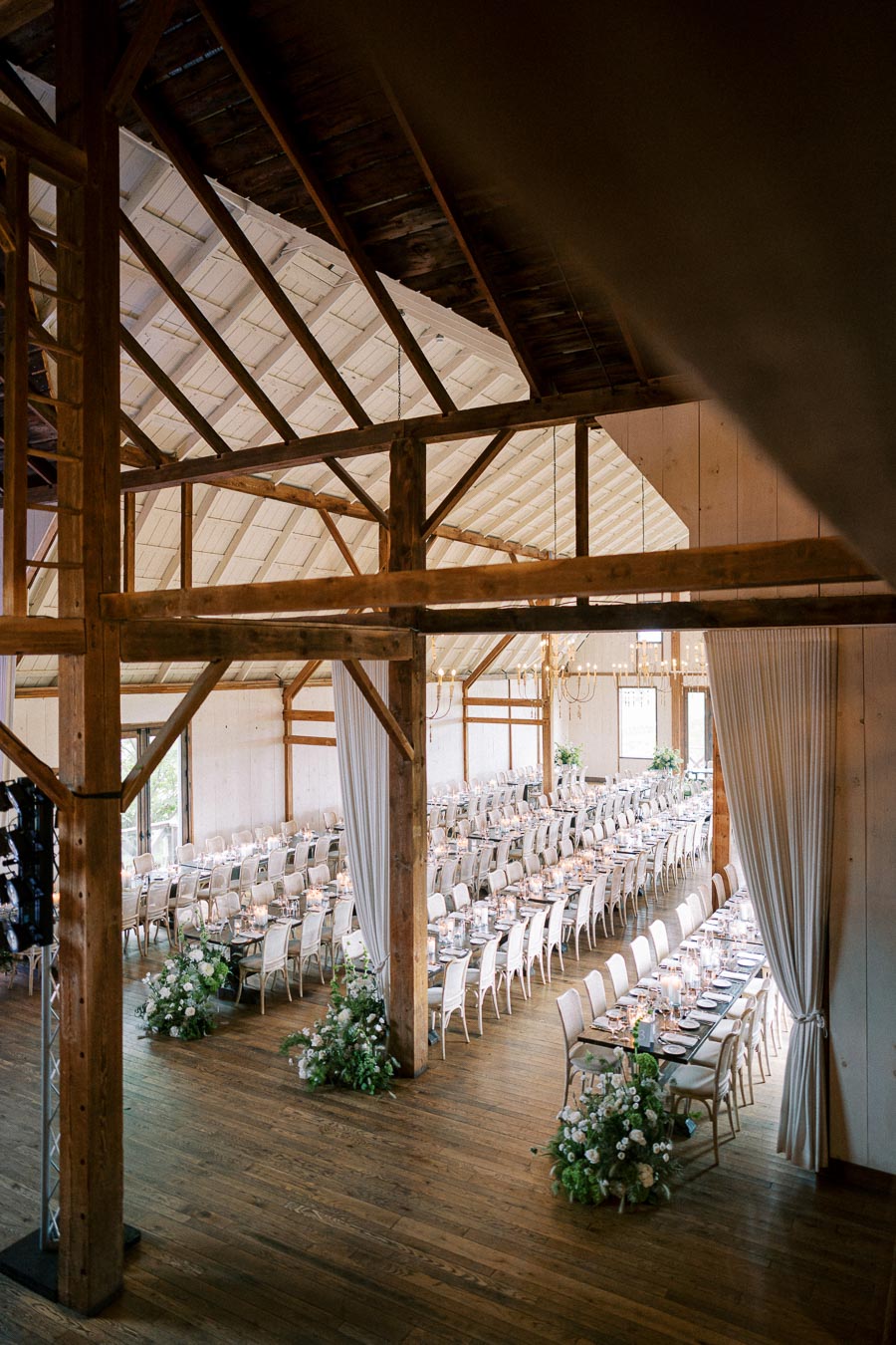Elegant rustic barn interior set for a wedding reception, featuring long banquet tables, white chairs, floral arrangements, and warm lighting, creating a charming and inviting atmosphere.