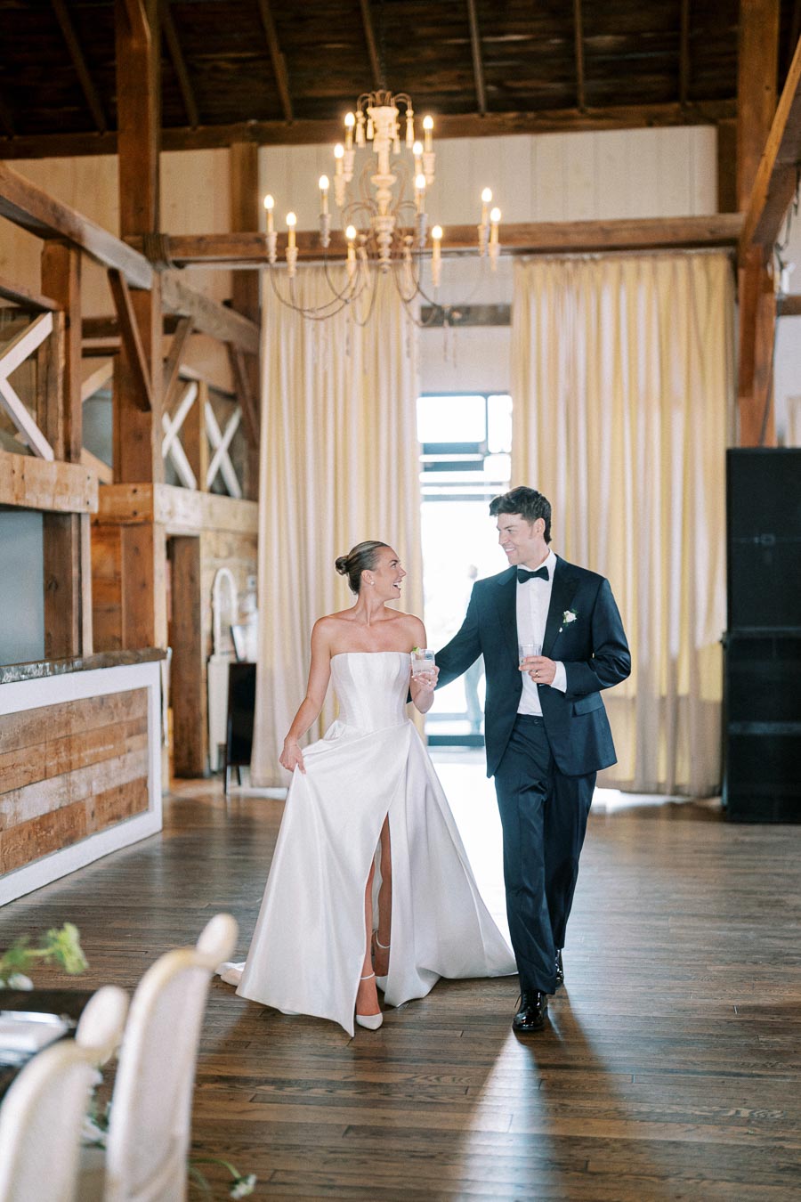 A bride in a strapless white gown with a thigh-high slit and a groom in a classic black tuxedo walk hand in hand, smiling, in a rustic wedding venue with wooden beams and a chandelier.