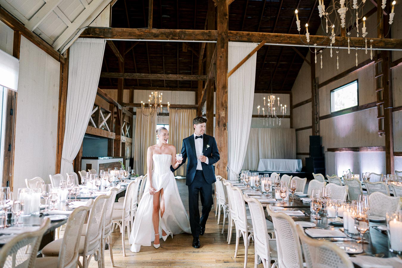 Bride and groom walk arm in arm through a rustic, elegantly decorated reception hall with white linens, chandeliers, and long dining tables adorned with candles and glassware.