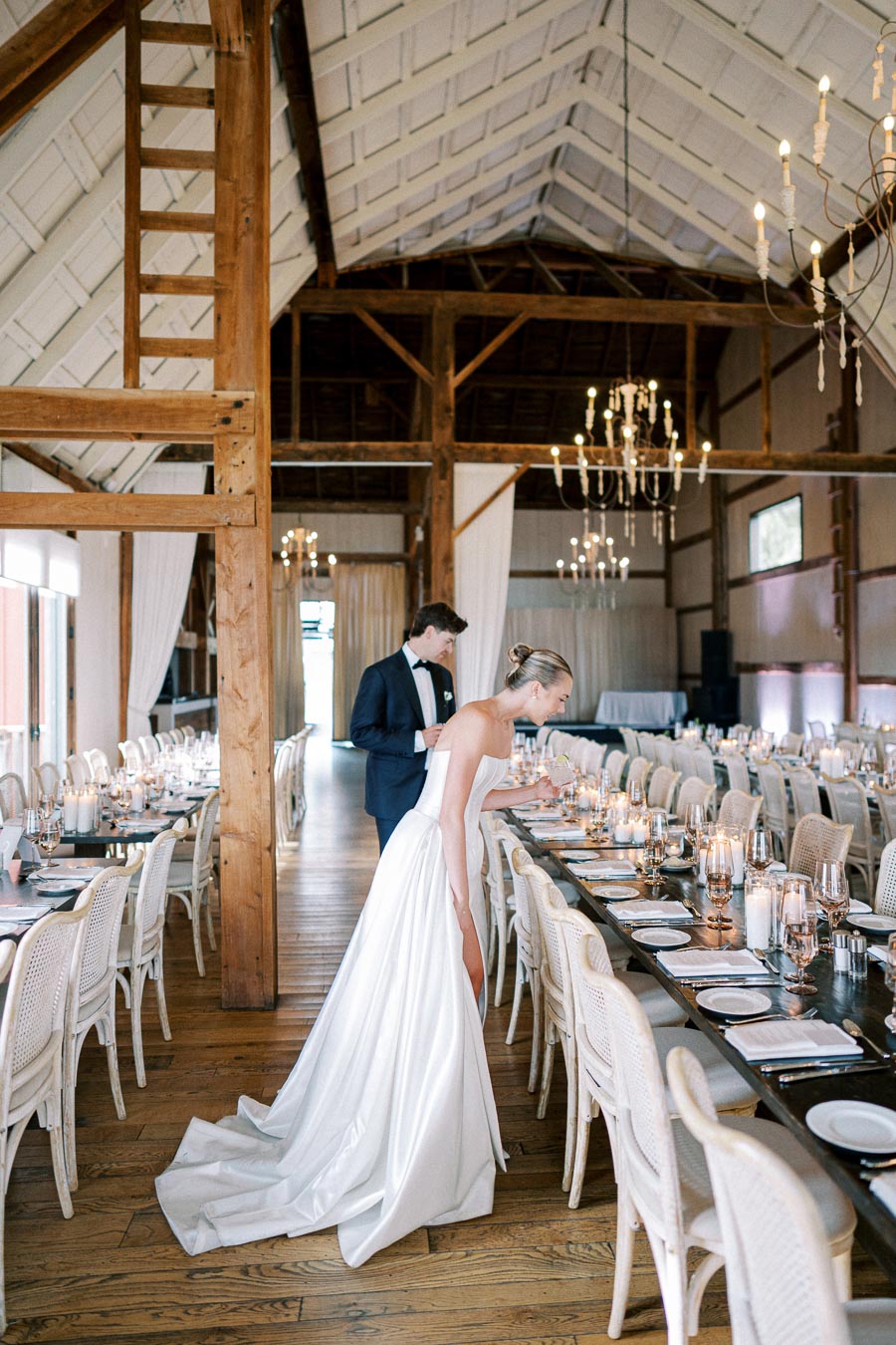 Bride and groom arranging elegant wedding reception table in a rustic barn venue, with wooden beams and chandeliers.
