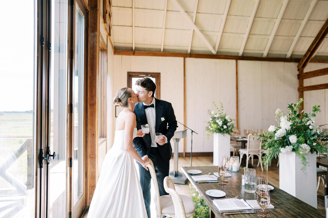 A bride and groom sharing a kiss at their elegant wedding reception with rustic decor, including lush floral arrangements and a wooden table setting.