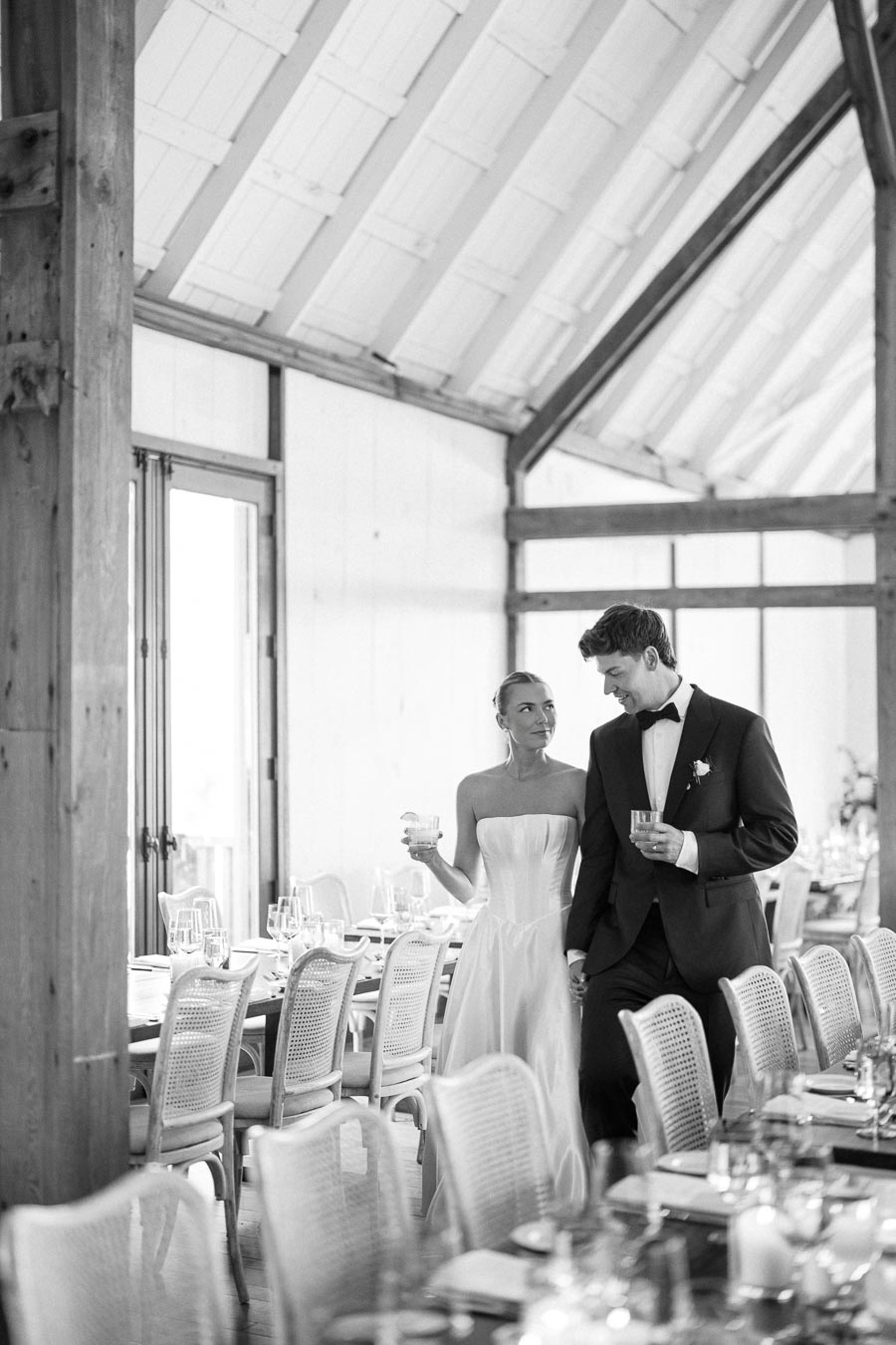 Black and white image of a couple in formal attire, holding drinks and standing in an elegant, wooden reception venue with a long table set for a celebration or wedding.